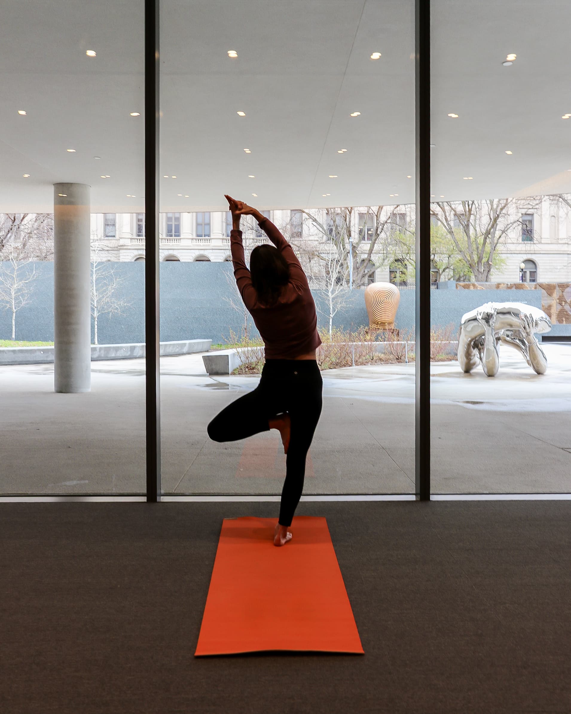 A person practices yoga on an orange mat indoors, standing in tree pose facing large floor-to-ceiling windows with a cityscape and outdoor sculptures visible outside.