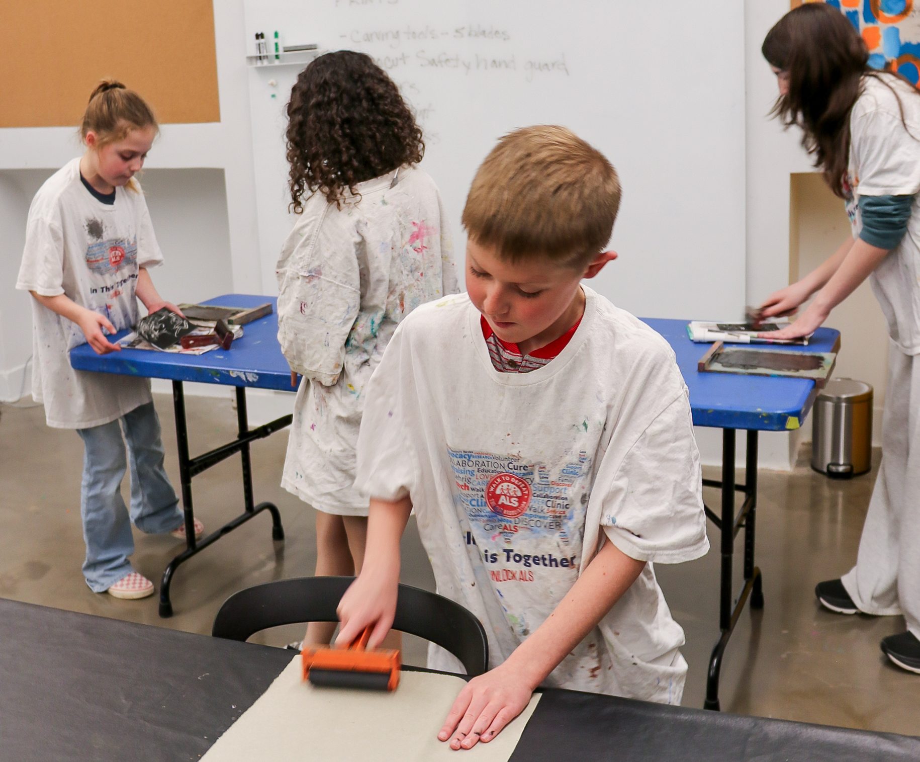 Four kids wearing white shirts work on art projects in a classroom. One boy in front uses a roller on paper, while three other kids work at a blue table in the background spreading ink.