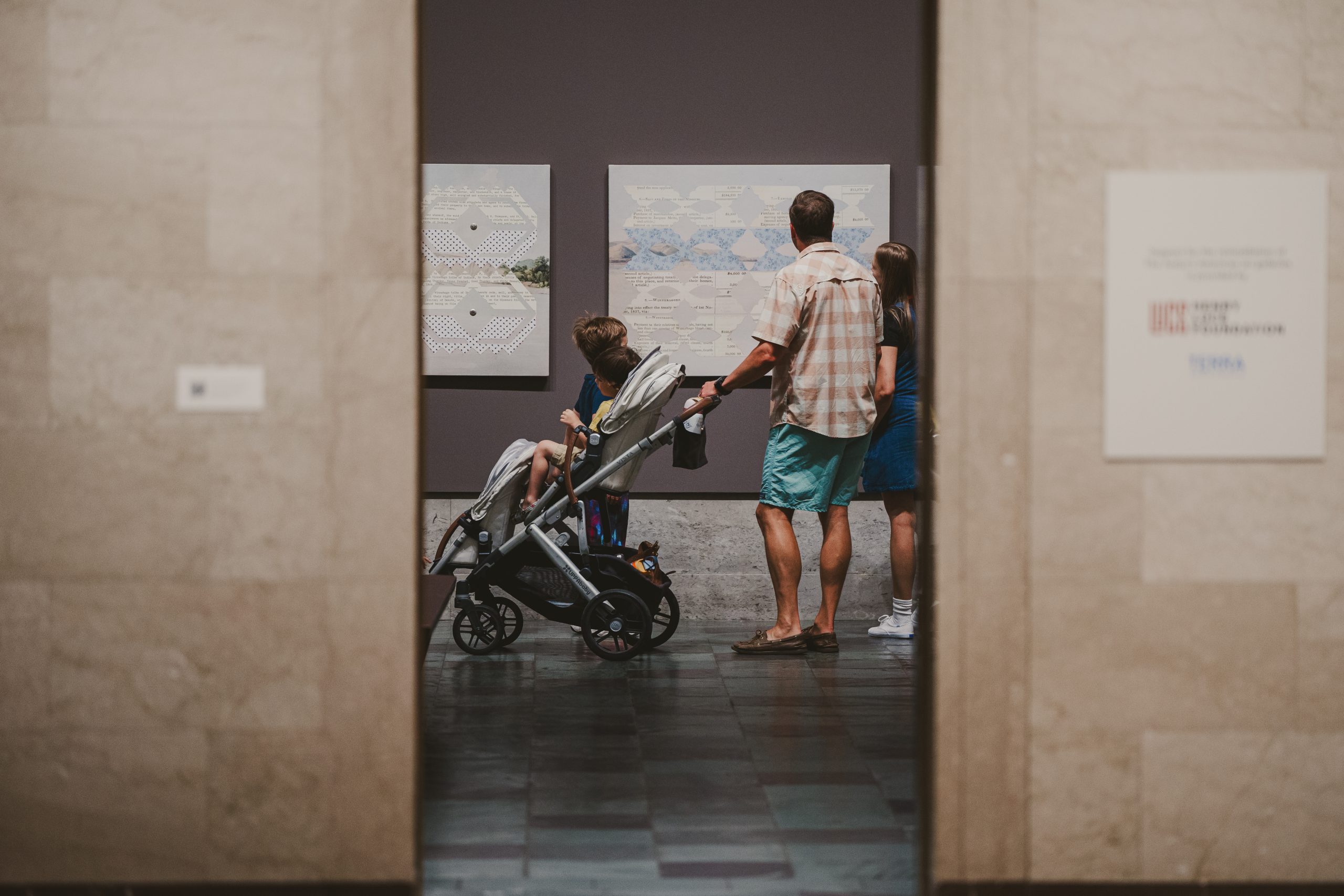 A family with a stroller looks at artwork on the wall of a museum gallery, viewed through a doorway. The adults and child are seen from behind, observing the exhibits.