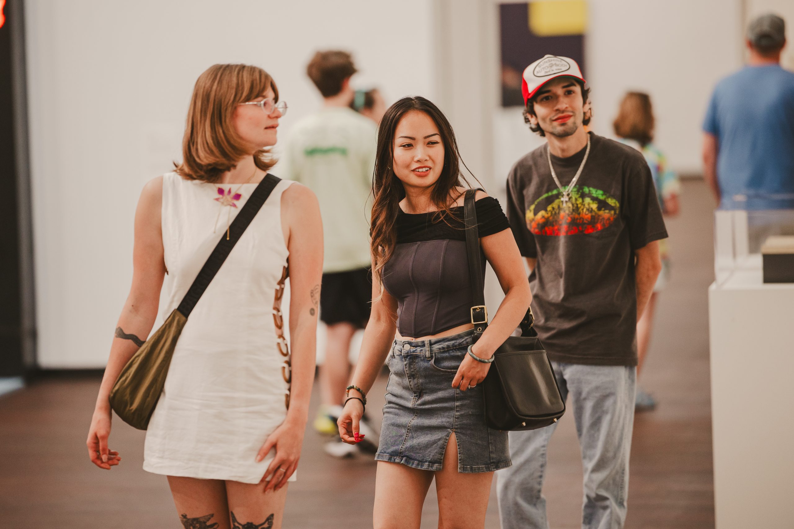 Three young adults walk together indoors. One wears a white dress, another a black top and denim skirt, and the third a graphic tee and cap. There are other people and artworks visible in the background.