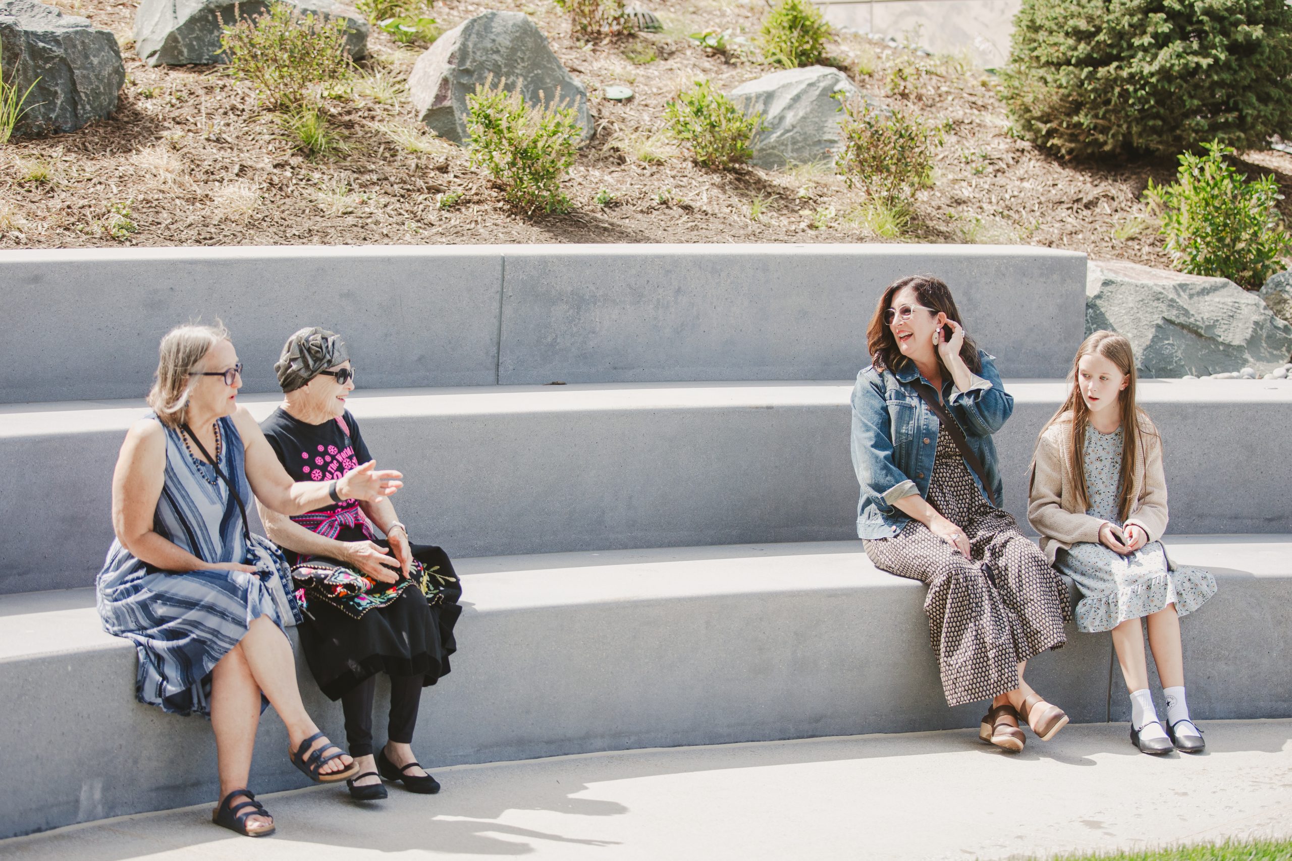 Four people sit on concrete amphitheater steps outdoors: two older women chatting on the left, and a woman with a young girl sitting together on the right. It's a sunny day with bushes and rocks behind them.