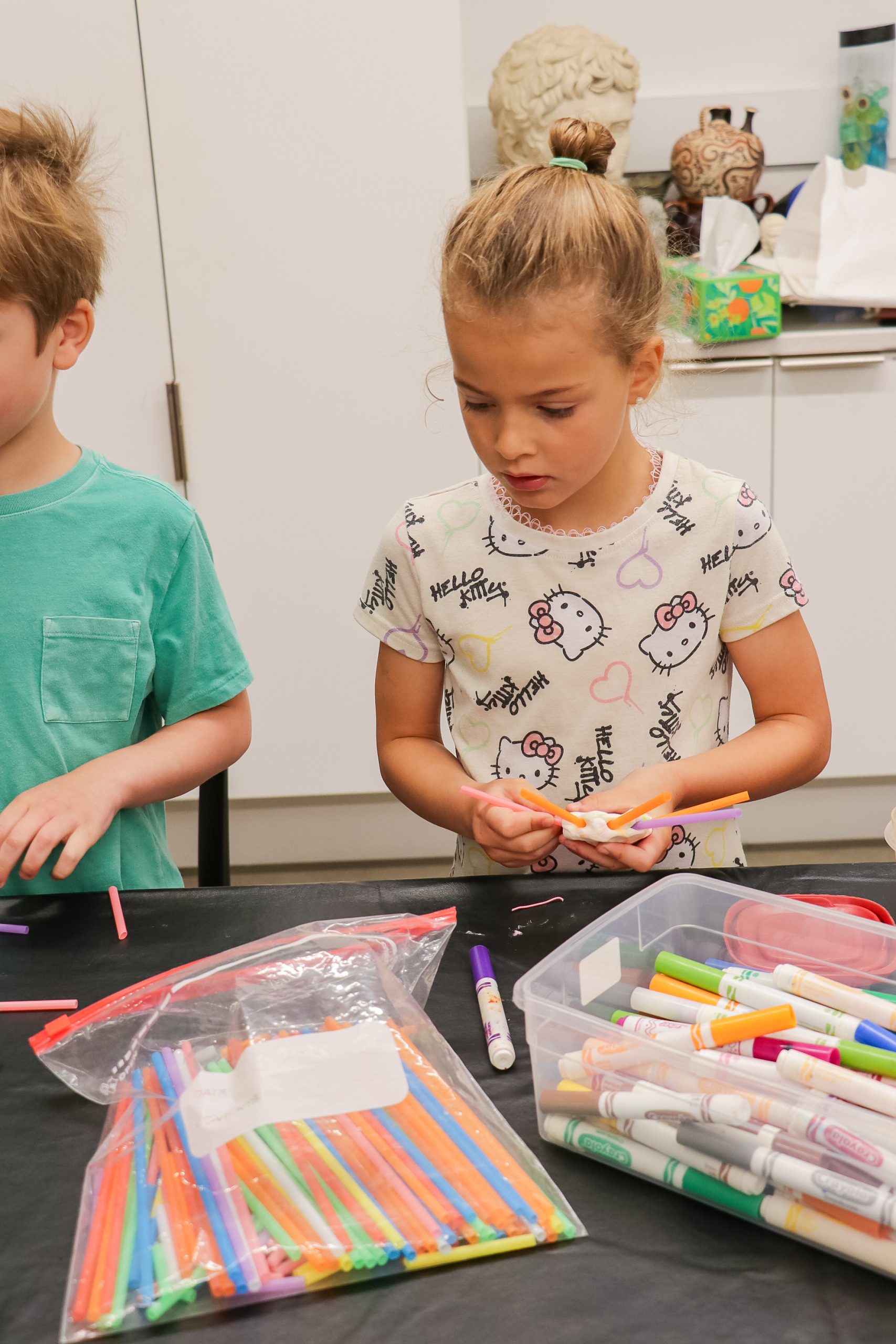 A young girl wearing a Hello Kitty shirt is selecting markers at a table covered with various markers and colored pens. Another child stands nearby, and the background shows a kitchen area.