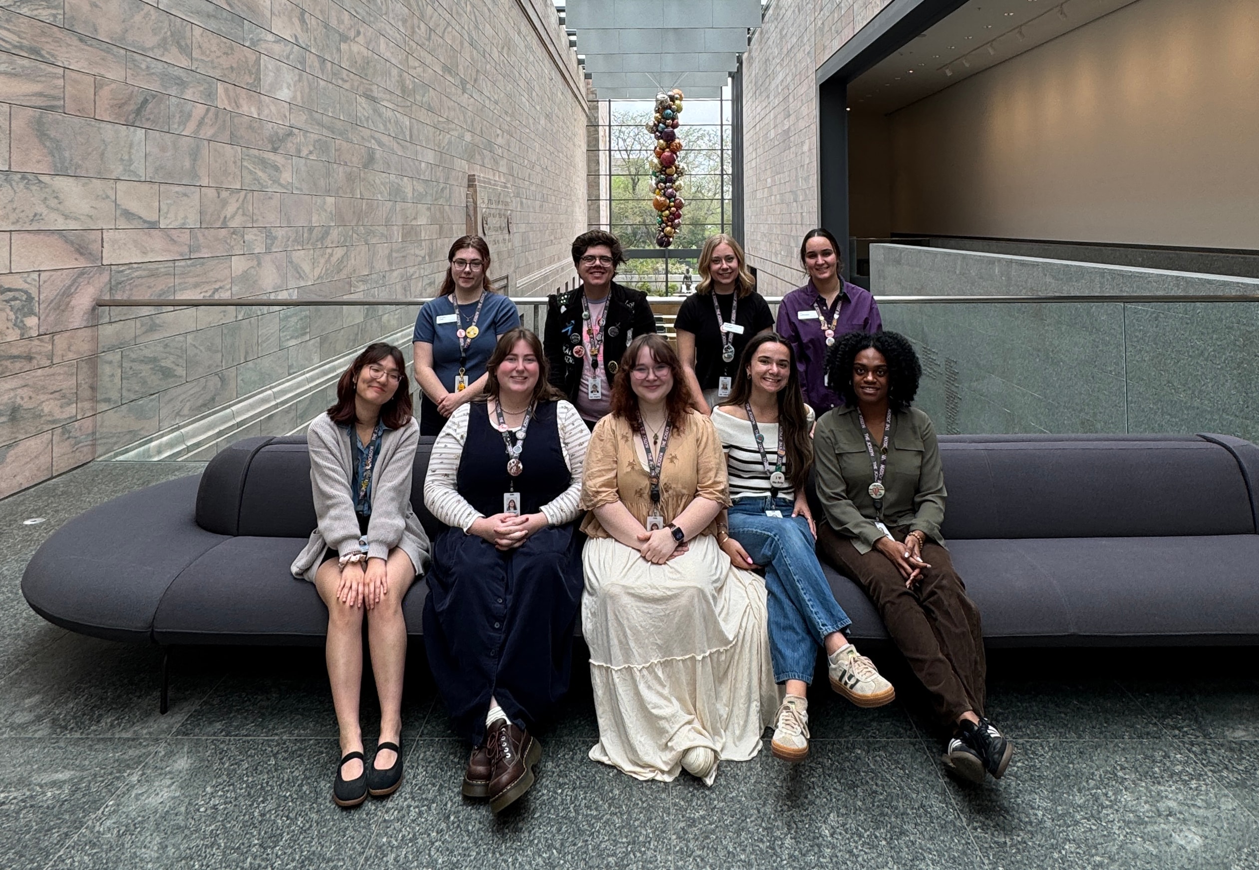 Nine people sit and stand together on and behind a modern gray couch in a spacious, well-lit gallery with stone walls and a tall glass window showing colorful hanging art in the background.