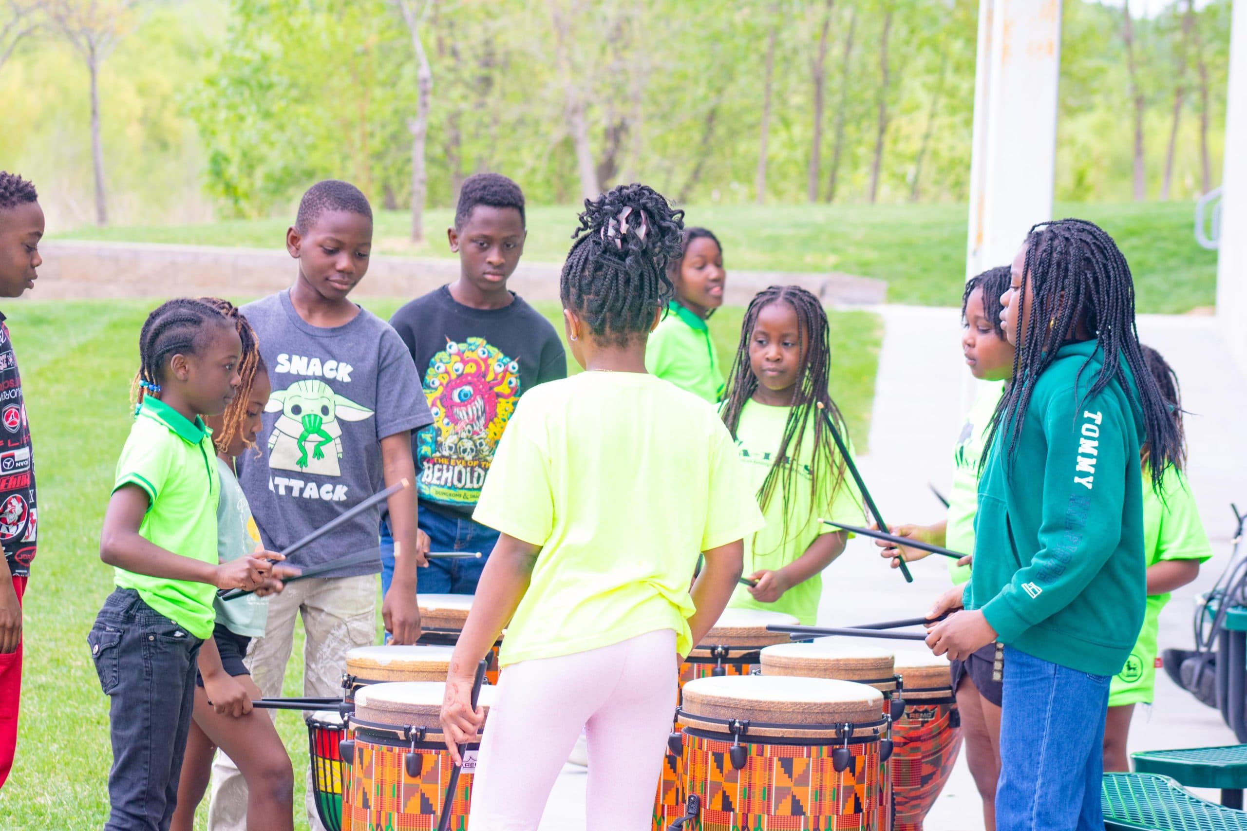 A group of children standing outdoors in a circle, playing drums together. Most are wearing bright green shirts, and the scene is set in a park with green trees in the background.