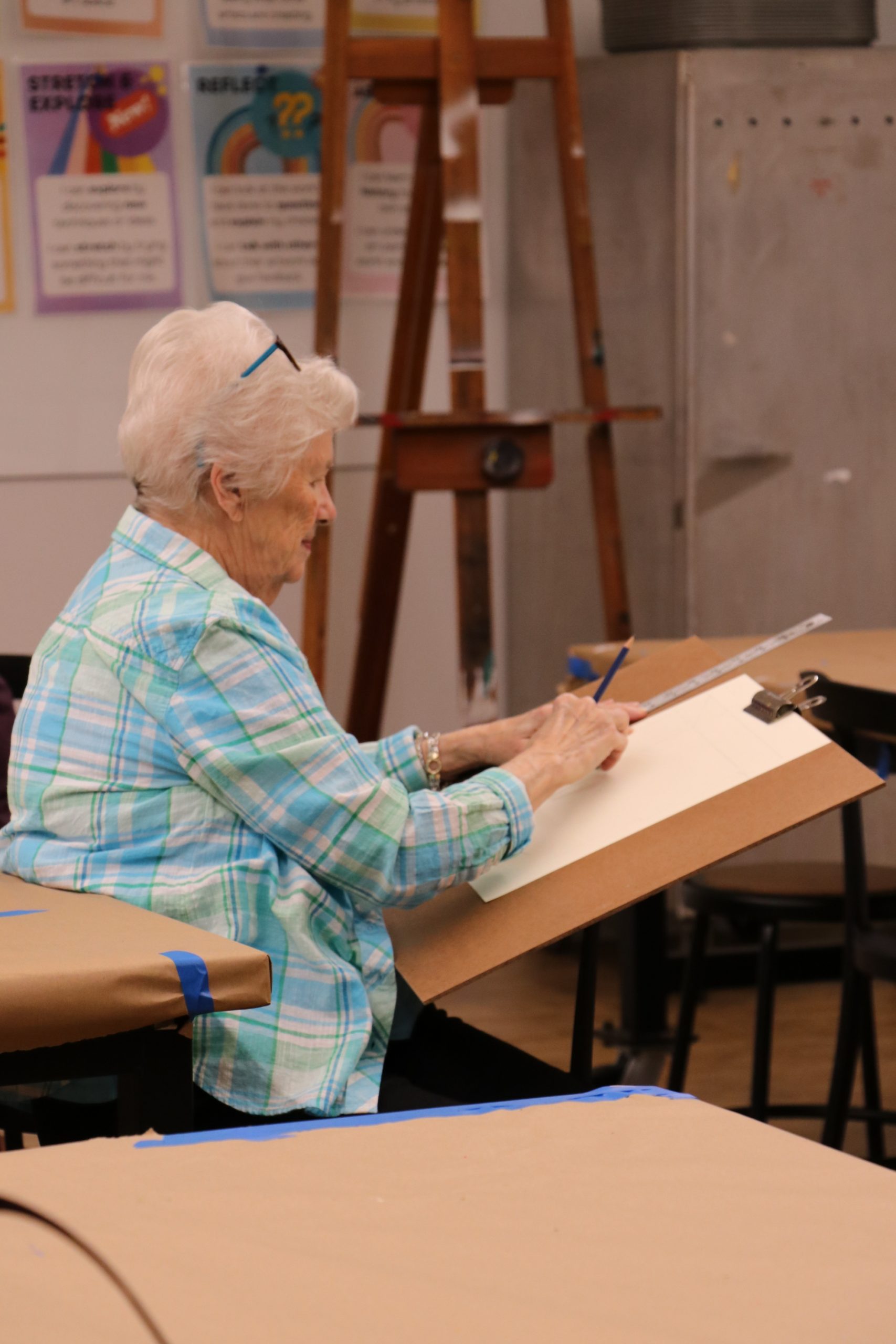 An older woman with white hair, wearing a blue plaid shirt, sits and sketches on a large clipboard in an art classroom. Art posters and an easel are visible in the background.