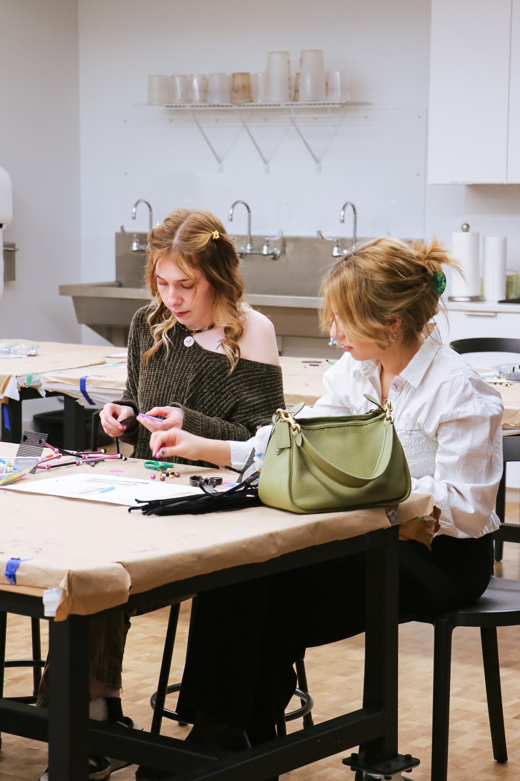 Two women sit at a table working on crafts or artwork in a studio, surrounded by art supplies. A green handbag rests on the table beside them. Shelves and a sink are visible in the background.