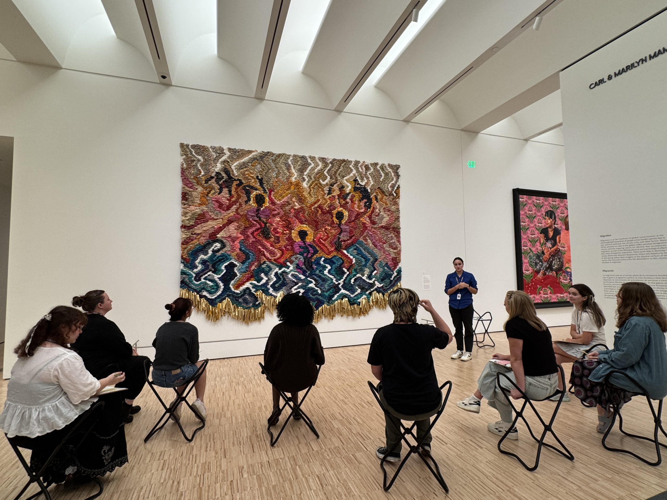 A group of people sit on foldable stools in an art gallery, listening to a guide speak in front of a large, colorful, textured wall hanging depicting abstract figures. White walls and natural light fill the modern space.