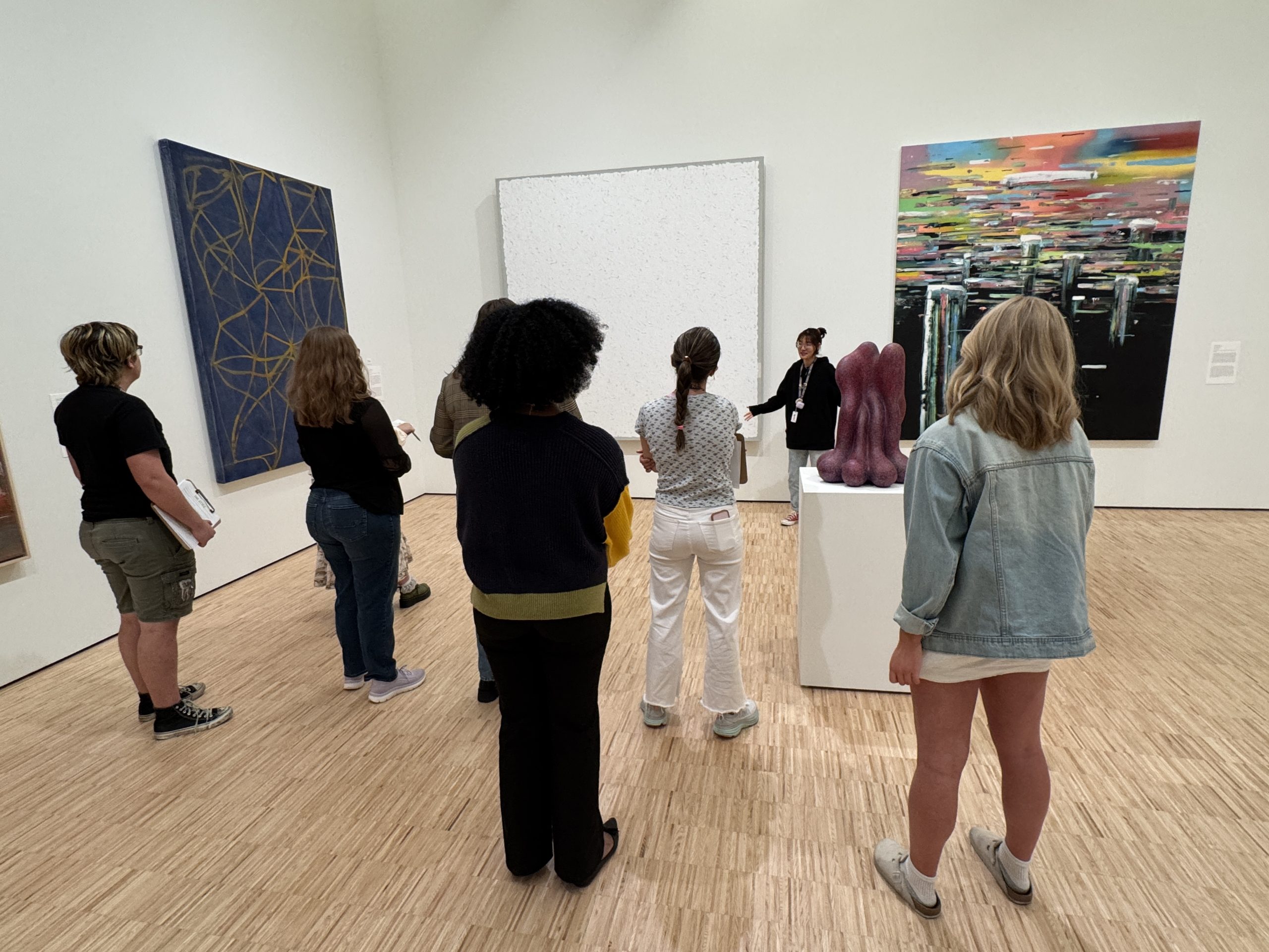 A group of seven people stands in an art gallery, listening to a guide speak in front of three large abstract paintings and a purple sculpture on a pedestal. The gallery has light wood floors and white walls.
