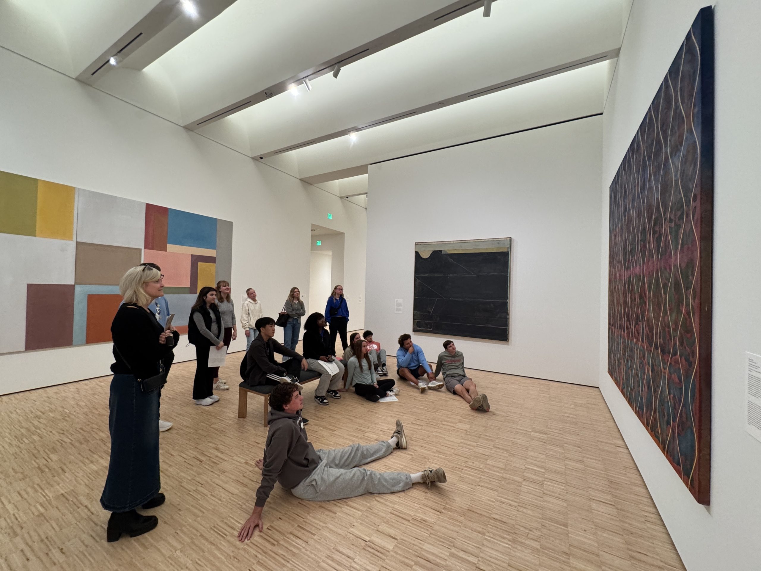 A group of people in a modern art gallery observe a large abstract painting on the wall; some are seated on benches or the floor, while others stand, all focused on the artwork.