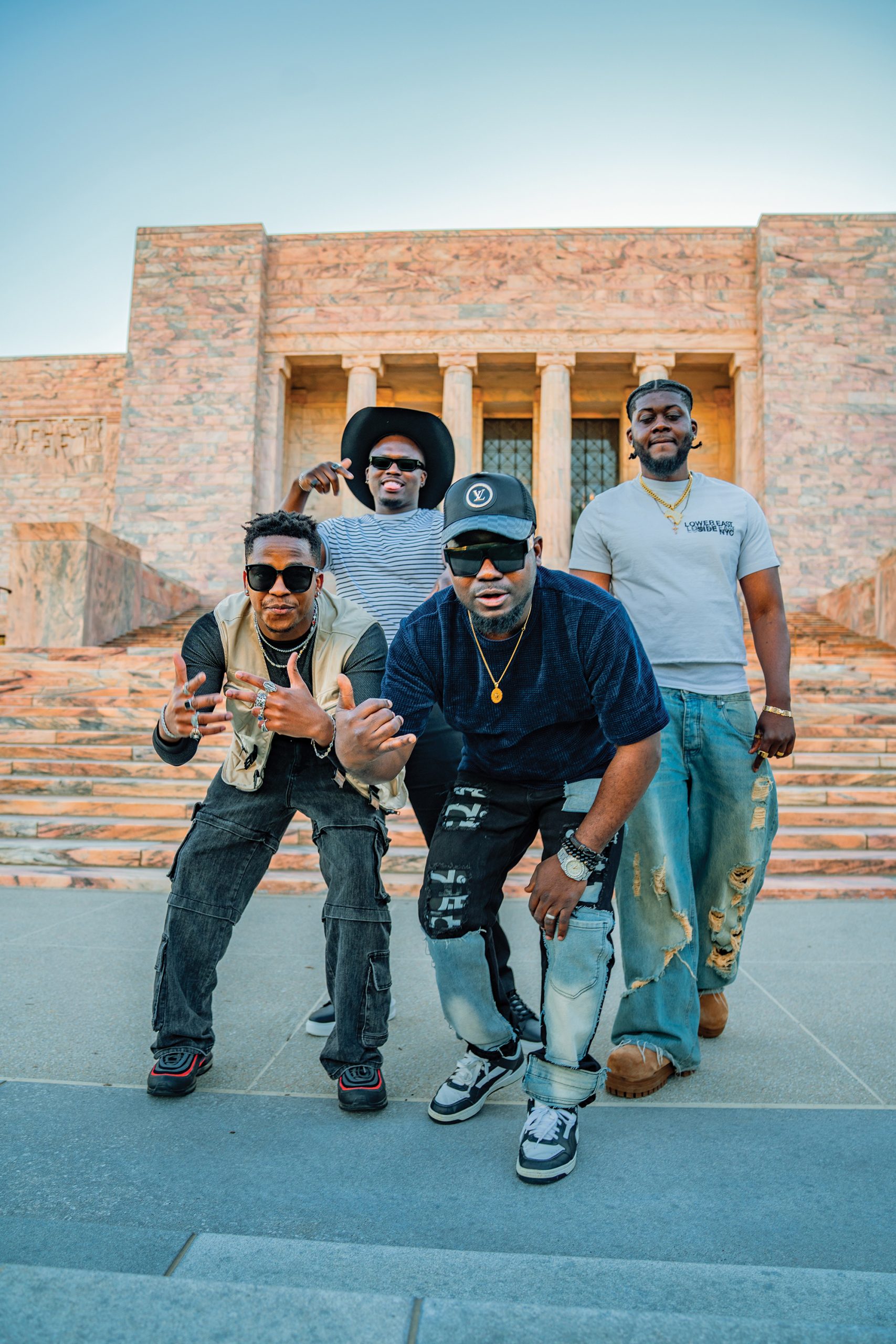 Four men pose on stone steps in front of a large building with columns. They wear casual, trendy outfits including hats, sunglasses, and jewelry, and appear to be having fun together outdoors.