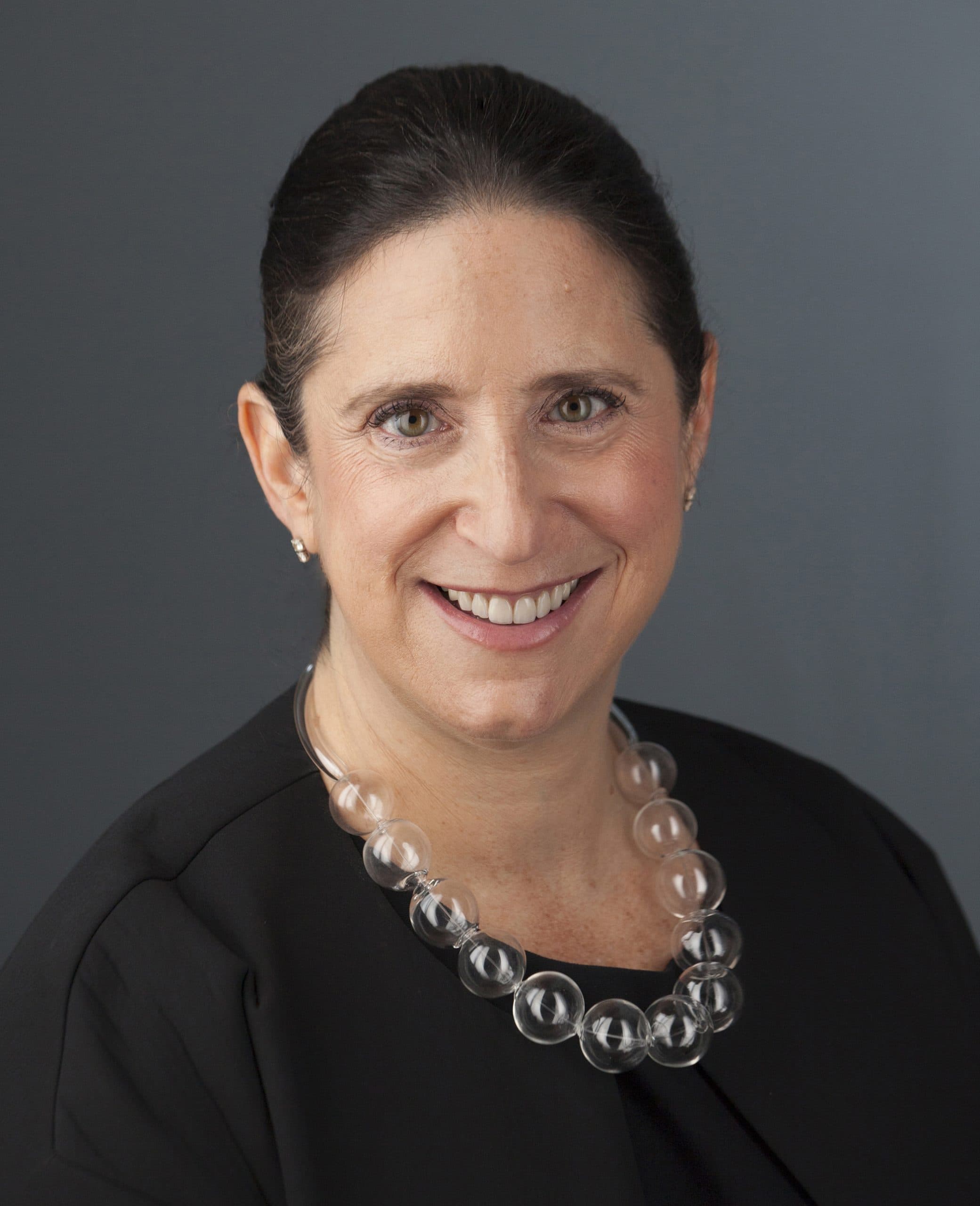A woman with dark hair pulled back wears a black top and a chunky, clear bead necklace, smiling in front of a plain gray background.