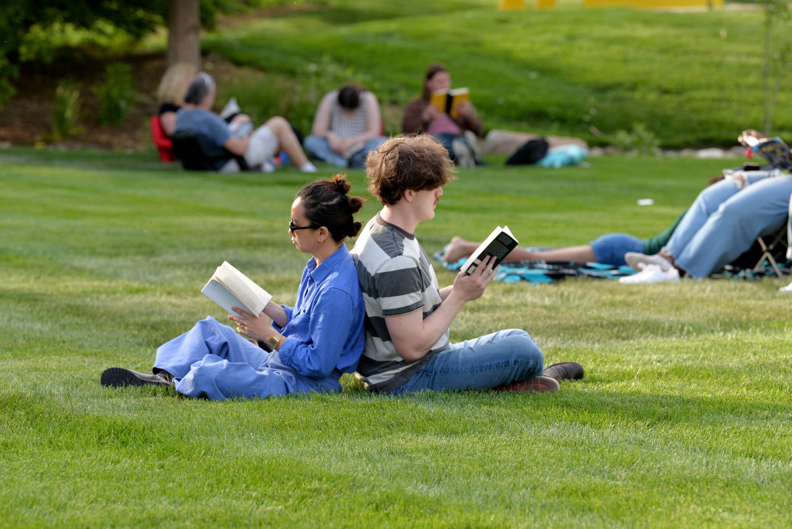 Two people sit back-to-back on green grass, each reading a book. Others are scattered around the lawn, also reading or relaxing in the background, creating a peaceful outdoor scene.