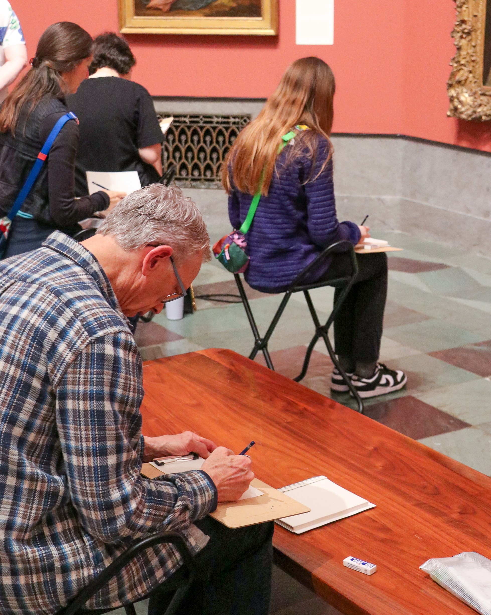 Several people sitting on stools in an art museum sketching in notebooks, focused on their drawings. The room has reddish walls, framed artwork, and a wooden bench in the foreground.