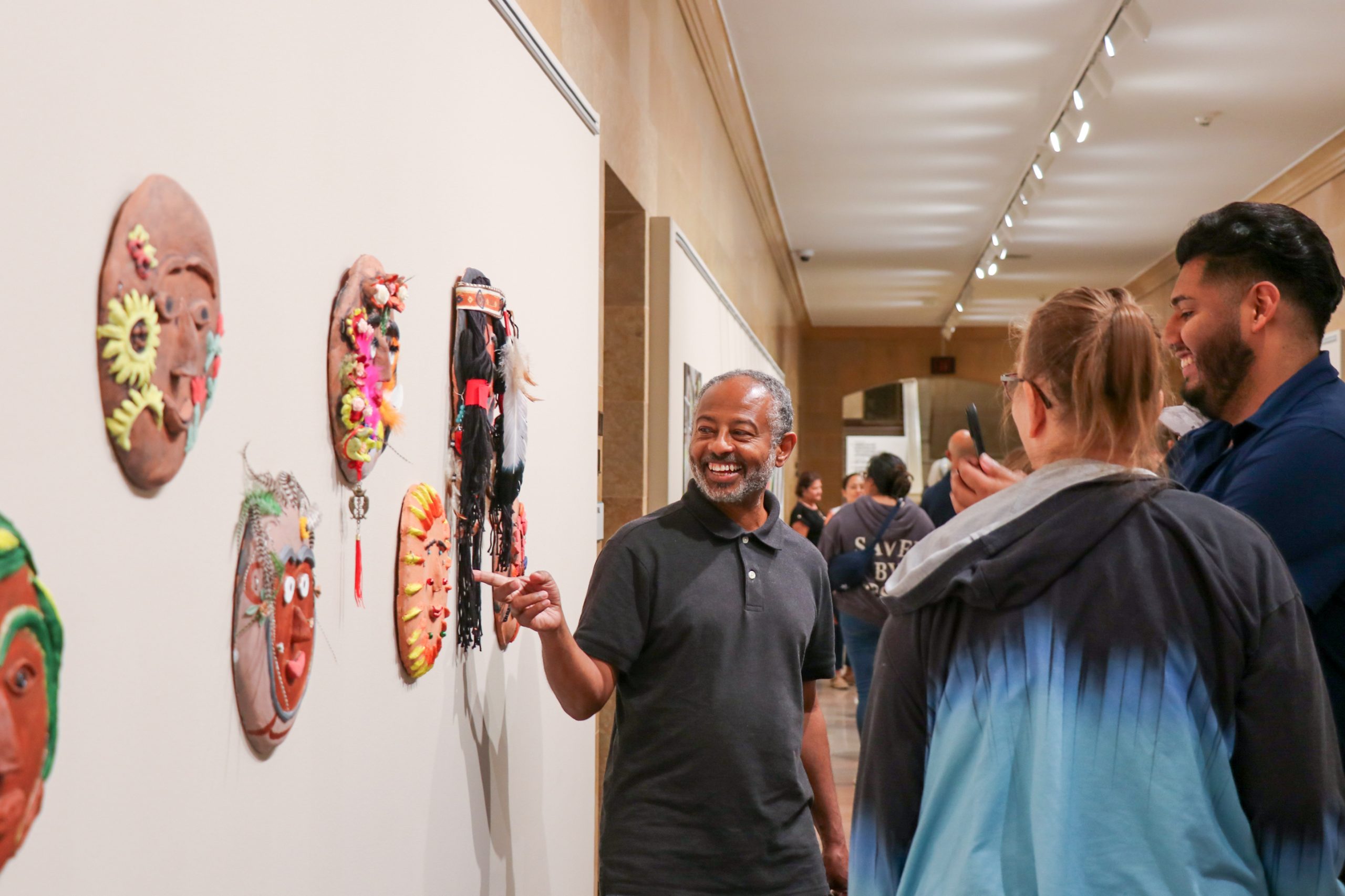 A smiling man points at colorful masks on a wall while talking to two people in a museum or gallery setting.