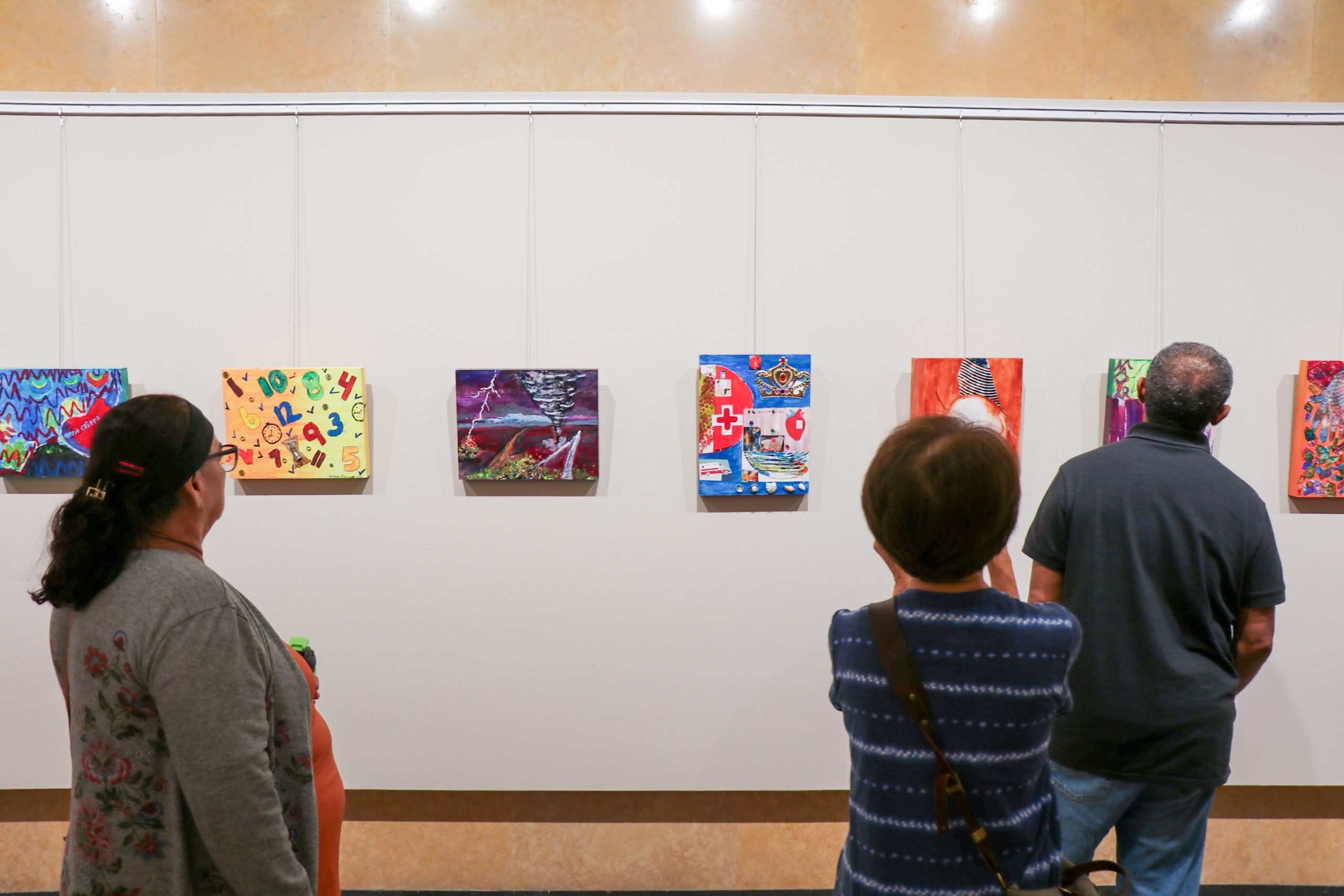 Three people look at colorful paintings hanging on a white wall in an art gallery. The abstract artworks are evenly spaced and small in size. The viewers are seen from behind.