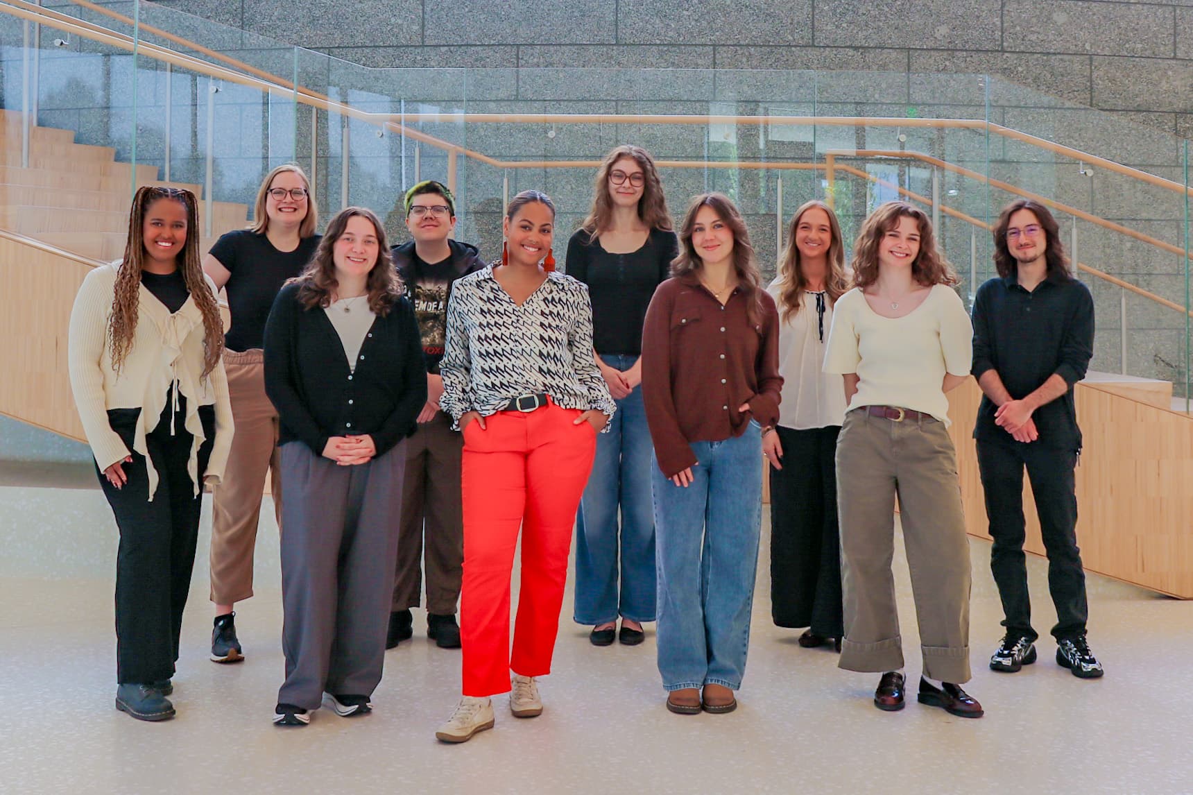 A group of eleven people stands indoors on a light-colored floor in front of stairs and a glass railing, smiling at the camera. The setting appears modern and brightly lit.