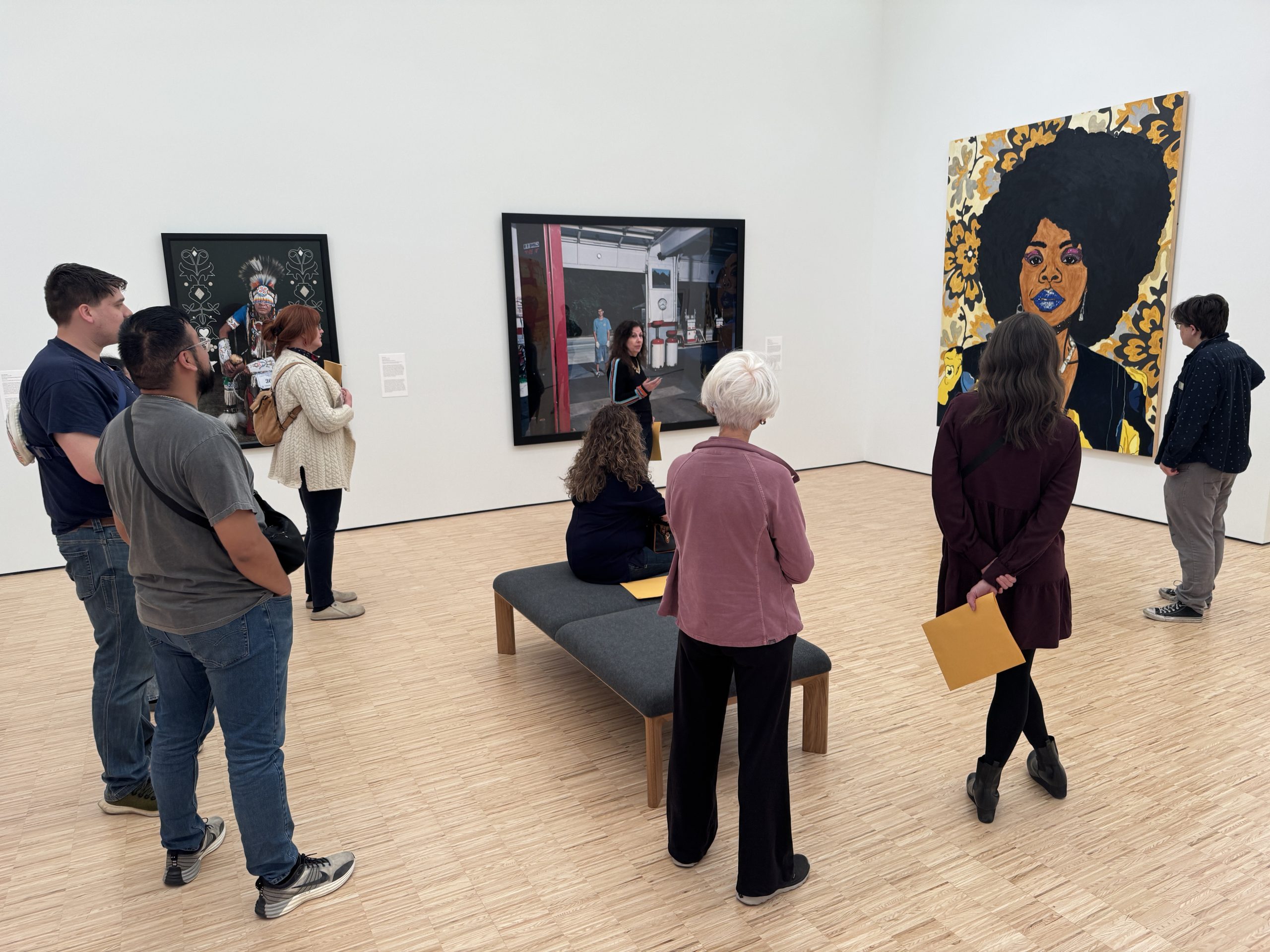 A group of people observes various artworks in a bright, modern gallery. One person sits on a bench while others stand. Art includes a large portrait of a woman with an afro on the right wall.