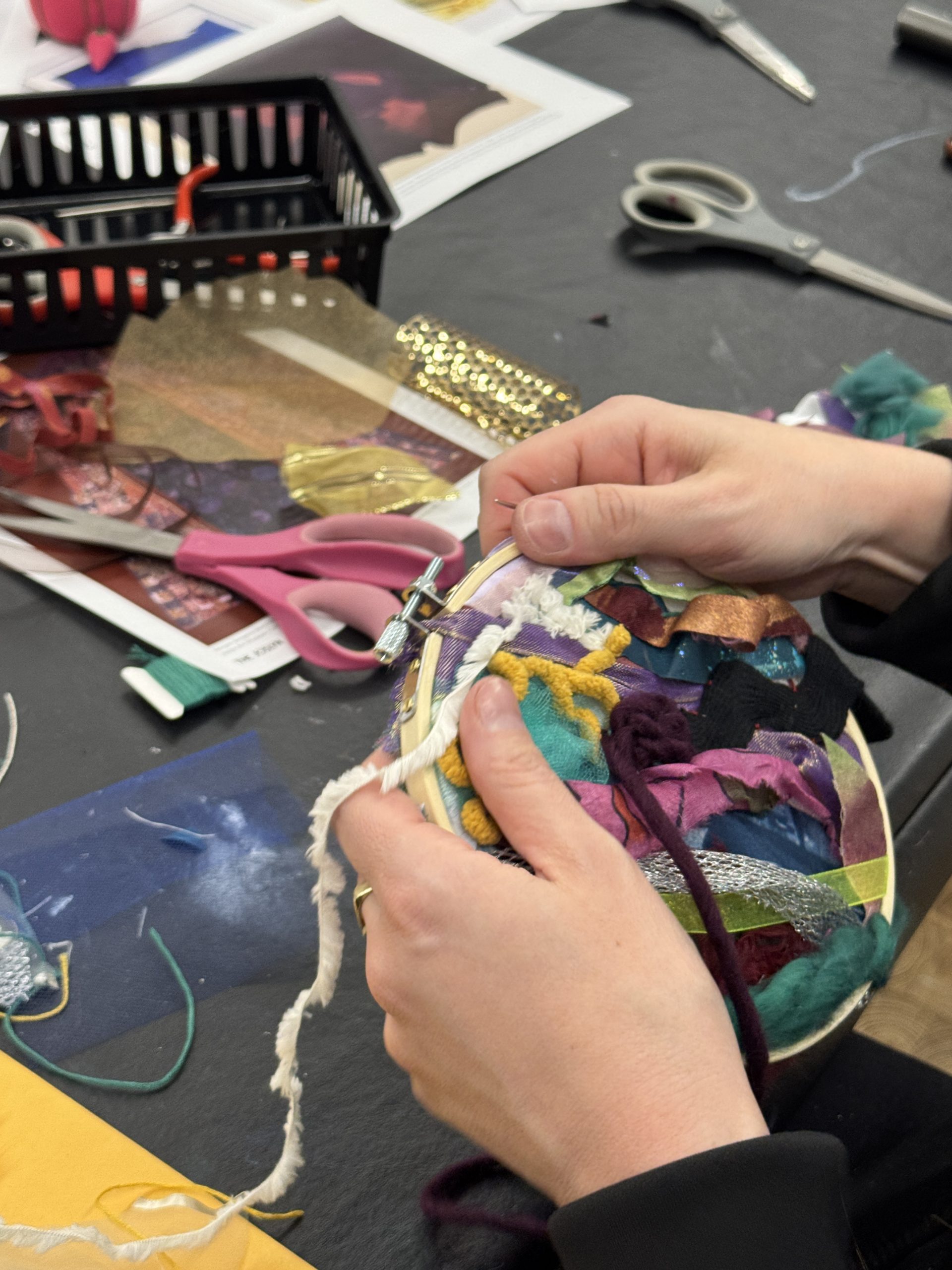 A person weaves colorful fabric strips onto a hoop, surrounded by scissors, ribbon, and art supplies on a black table.