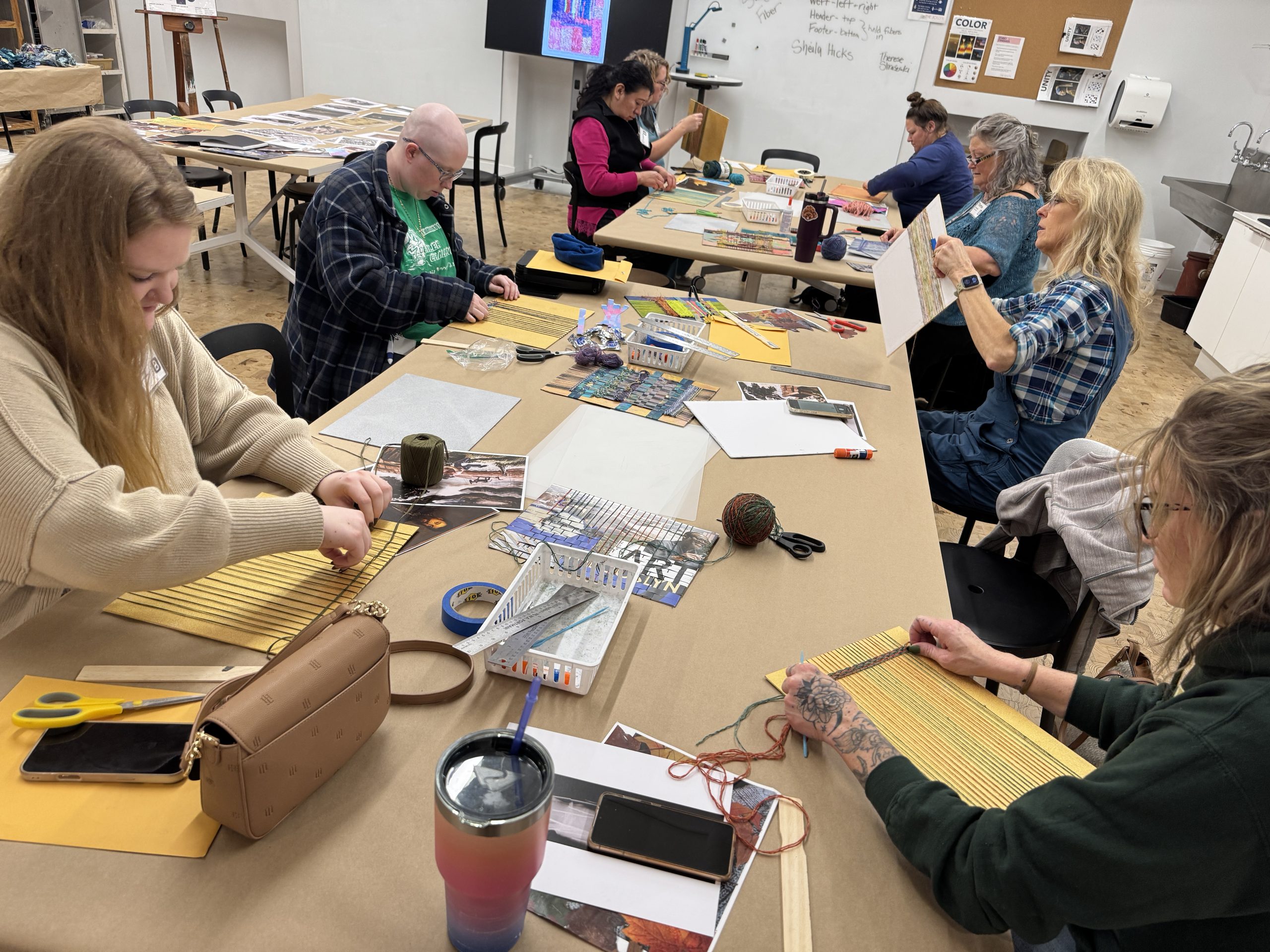 A group of people sit around a table in an art classroom, working on weaving projects with yarn, paper, and other materials. Art supplies, cups, and purses are scattered on the table.