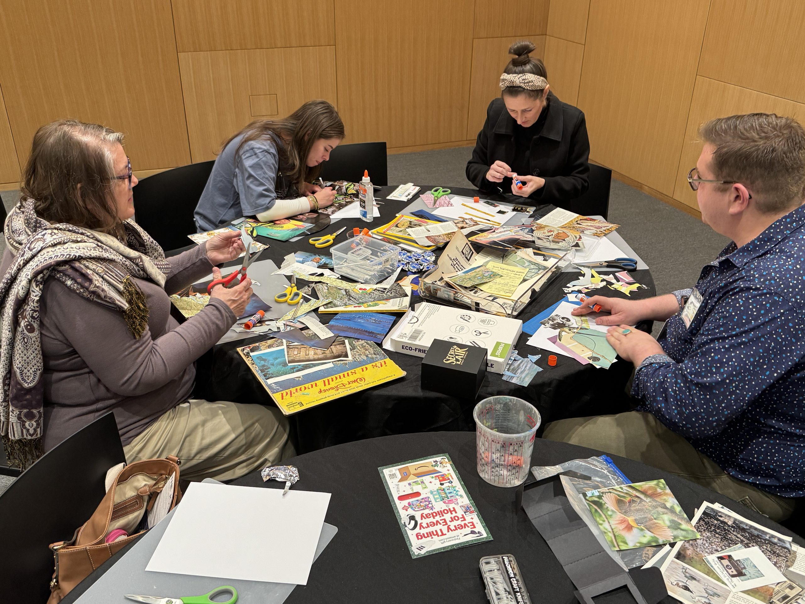 Four people sit around a table covered with art supplies, magazines, scissors, and glue, creating collages together in a well-lit room with wooden walls.
