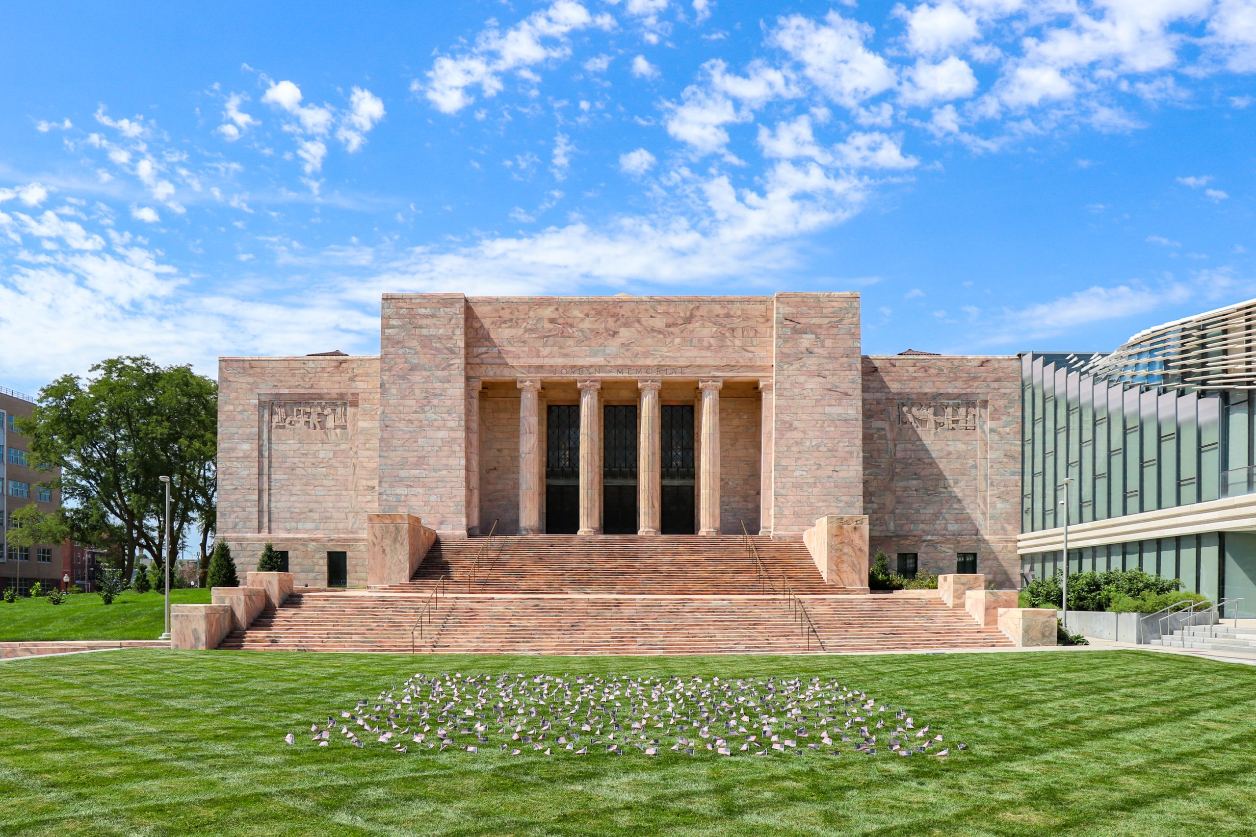 A large, symmetrical stone building with tall columns and wide steps sits under a blue sky, surrounded by green lawn and modern glass buildings on either side.