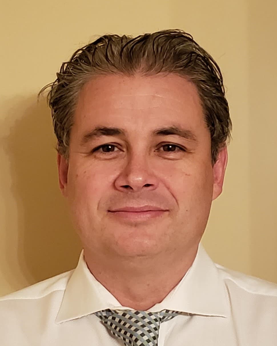A man with short brown hair, wearing a white dress shirt and a patterned tie, stands in front of a beige background, looking at the camera and smiling slightly.