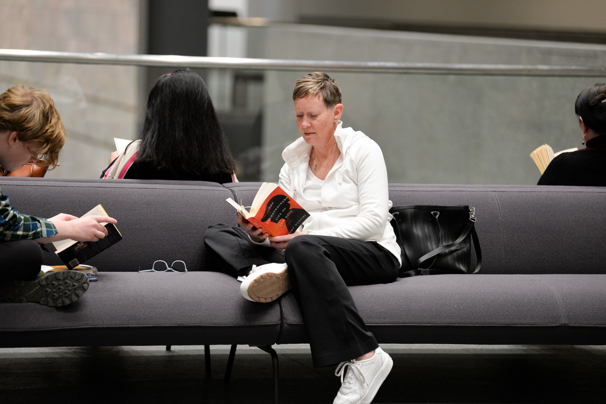 A person in a white shirt sits on a gray couch reading a book, with others nearby also reading. A black bag sits next to them on the couch.
