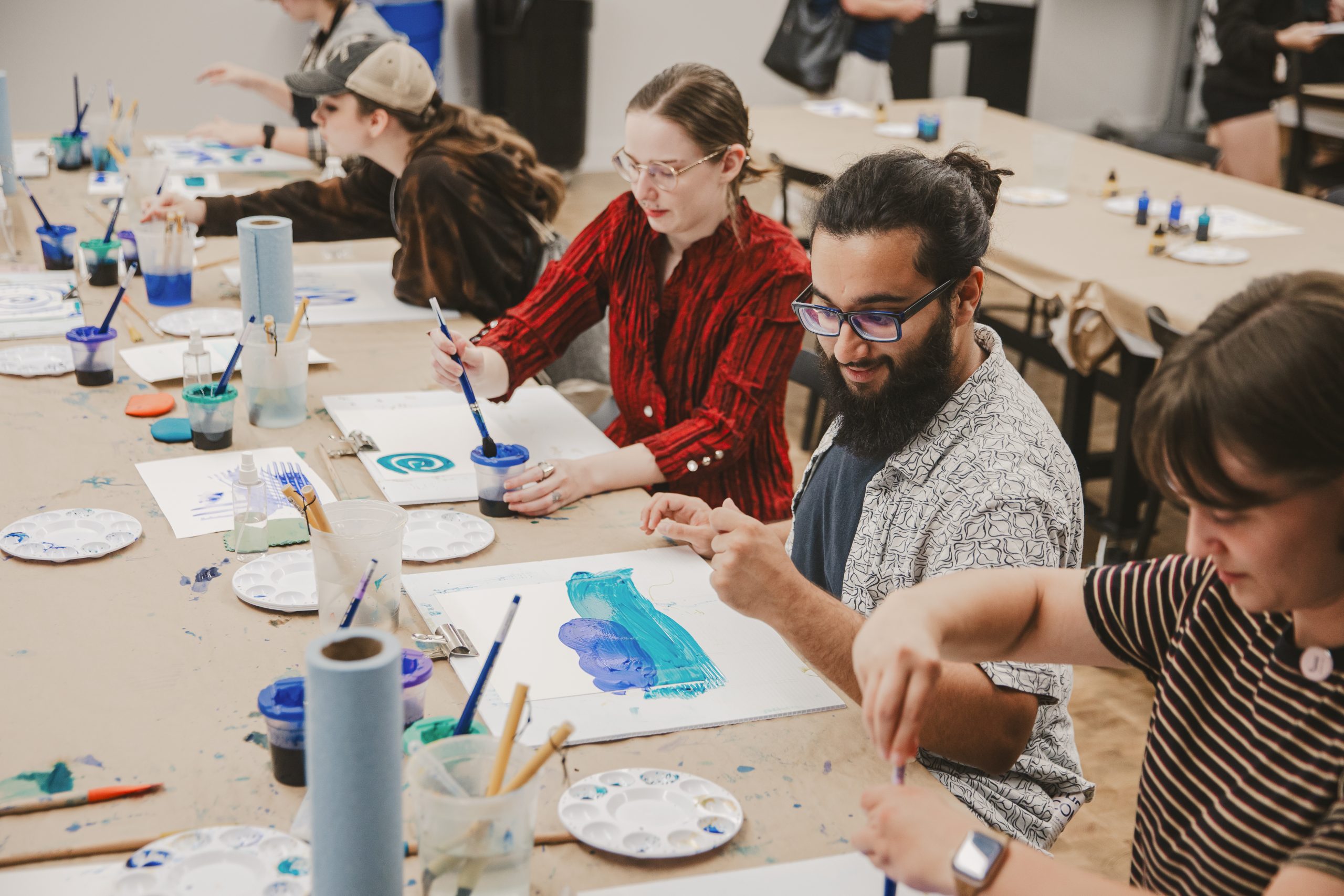 A group of people sit at a long table painting with blue and green watercolors. Art supplies, palettes, and cups are scattered on the table. The atmosphere appears creative and focused.