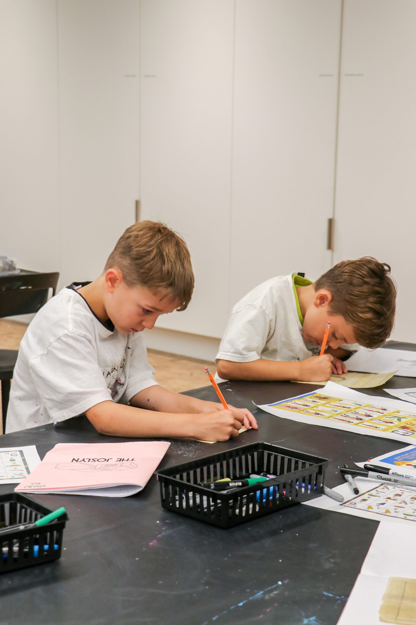 Two young boys sit at a table, focused on drawing or coloring with pencils. Art supplies and papers are spread out in front of them in a classroom setting.
