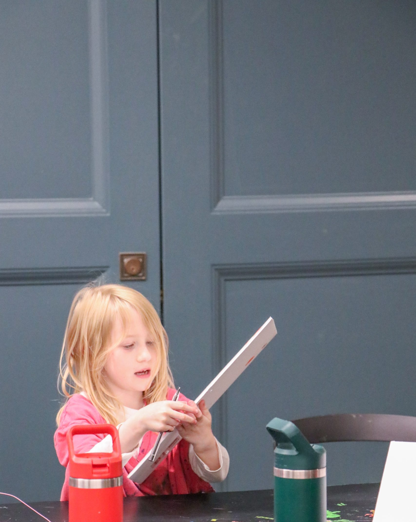 A young child with blonde hair holds a craft project at a table with two water bottles and a name card reading “Clara B.” The background is a blue door.