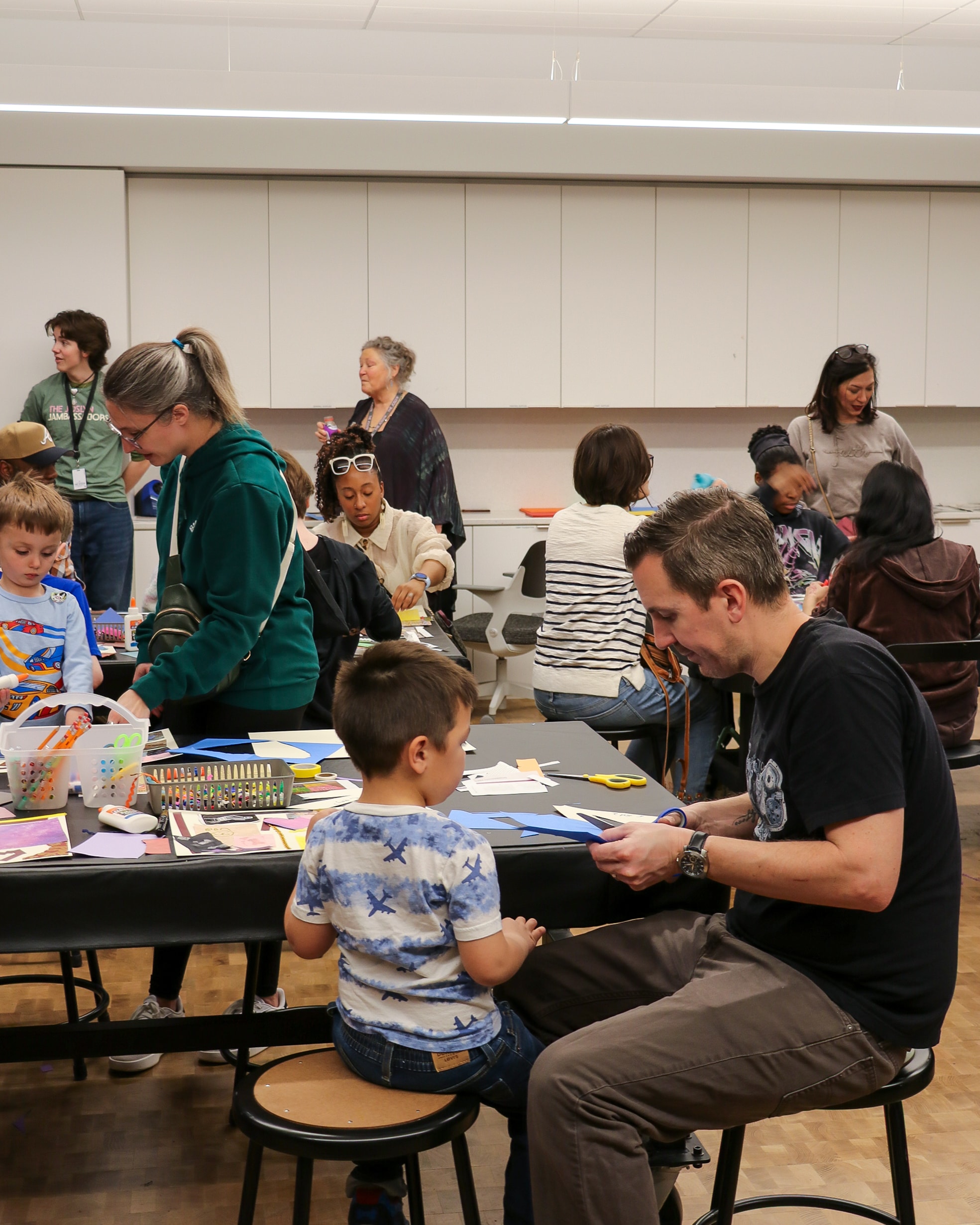 Adults and children are sitting and standing around tables in a classroom, working on arts and crafts activities with colored paper and supplies.