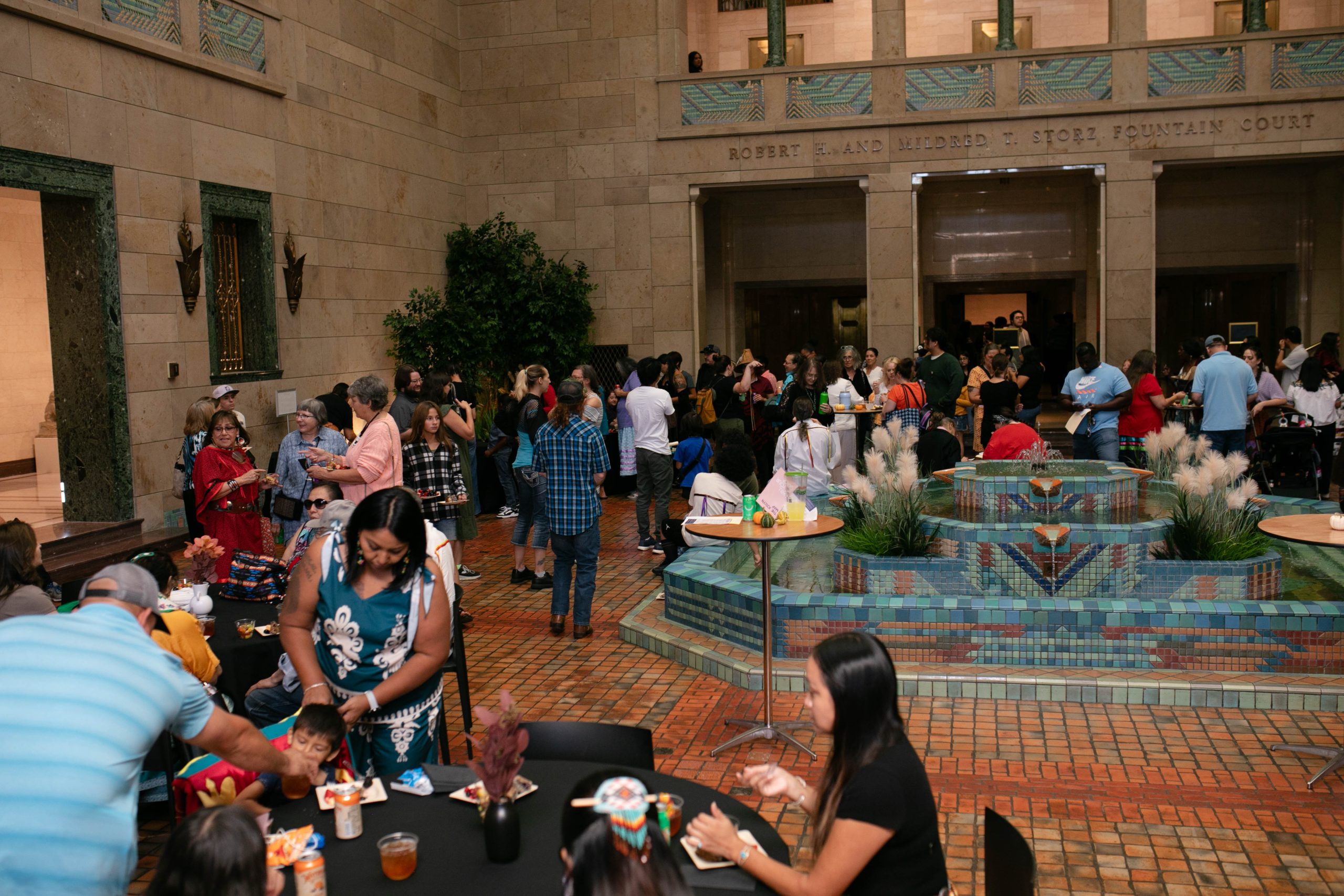 A large group of people gather indoors around tables with food and drinks, near a decorative tiled fountain in a spacious, high-ceilinged hall with stone walls and greenery.