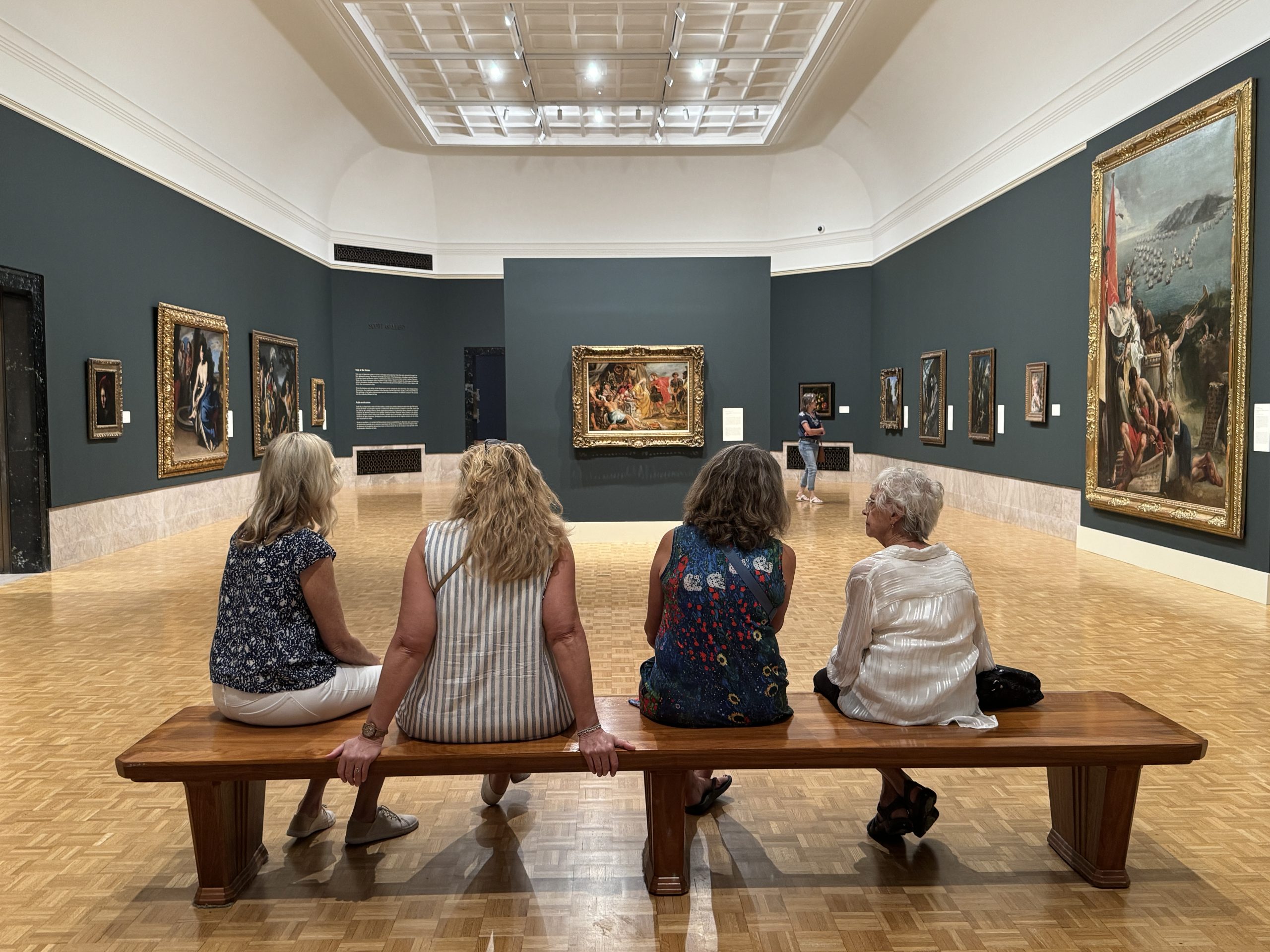 Four women sit on a wooden bench in an art gallery, facing classic paintings displayed on dark green walls. The room has a parquet floor and a high, bright ceiling. Another visitor sits alone in the distance.