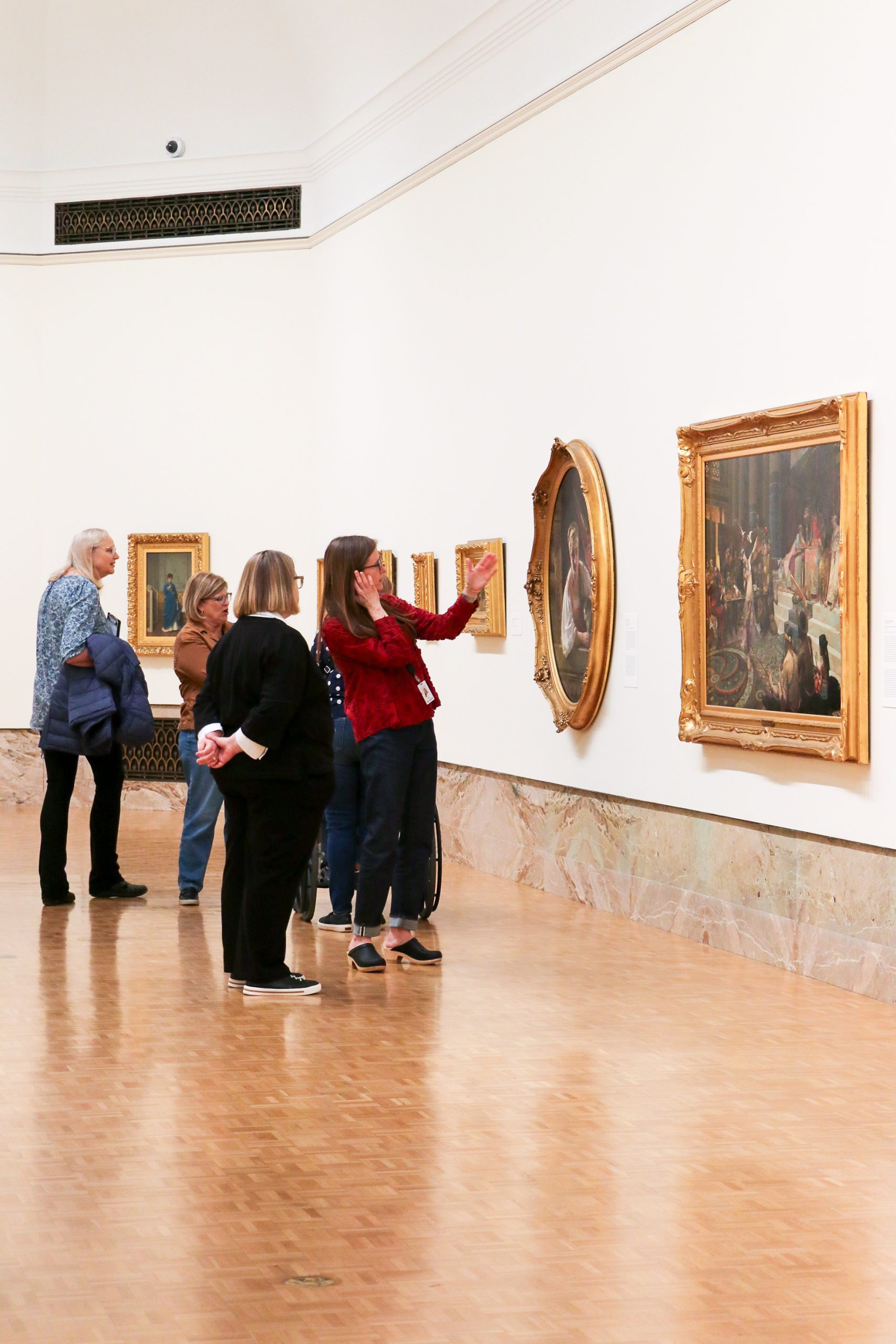 A group of people stands in an art gallery, viewing and discussing paintings displayed on a white wall. The floor is wooden, and the scene appears calm and contemplative.
