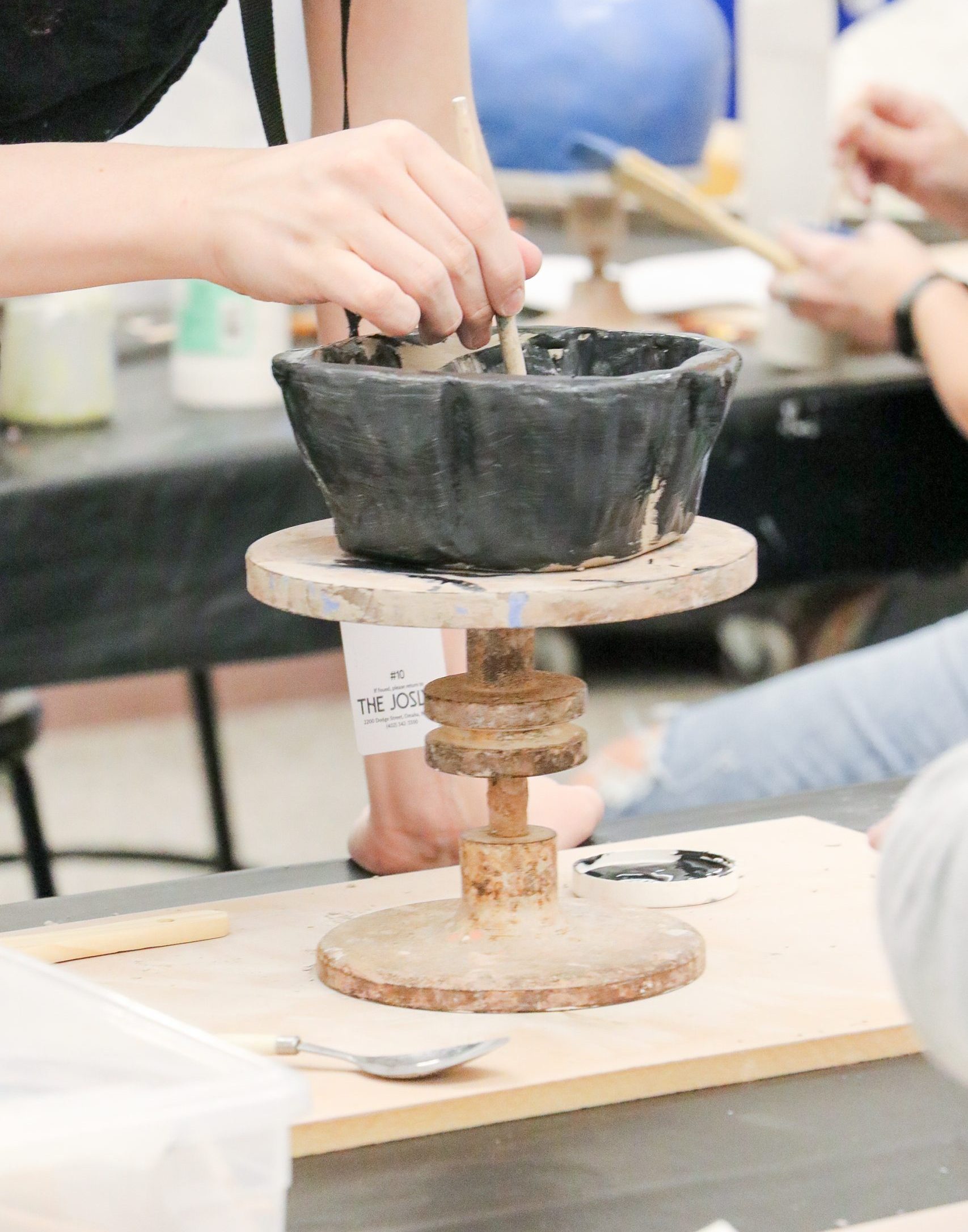 A person uses a brush to paint a black ceramic bowl on a spinning wheel during a pottery class. Another person sits in the background, partially visible.