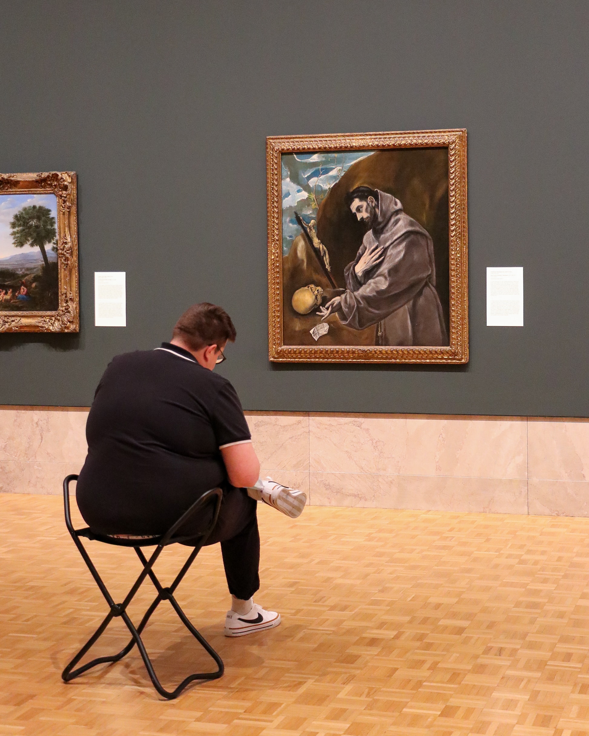 A person sits on a folding stool in front of a framed painting in an art museum, looking at the artwork on the wall. The gallery features wooden floors and green walls.