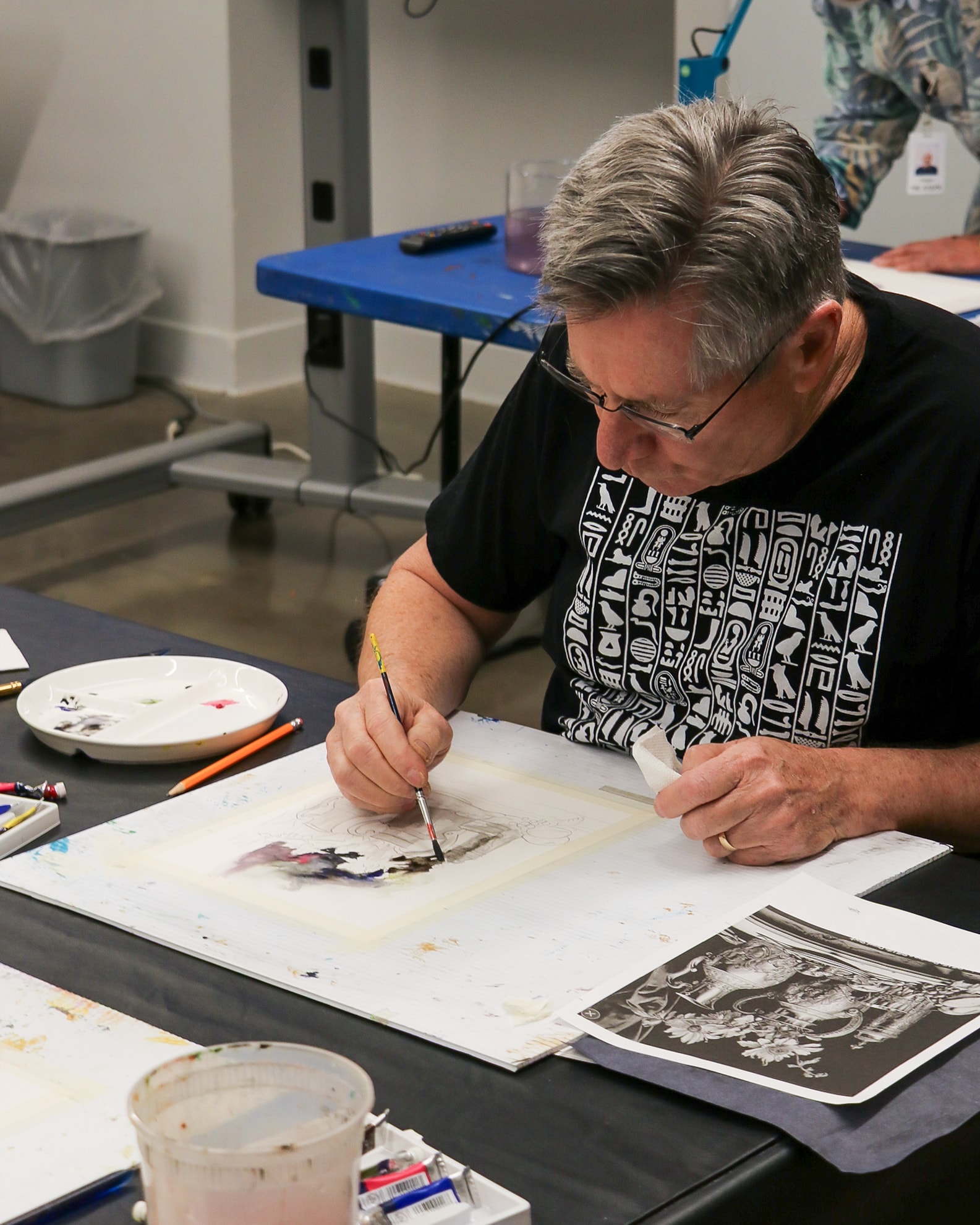 An older man with gray hair and glasses sits at a table, painting on paper with watercolors. Art supplies and reference images are spread out on the table around him in a bright studio setting.