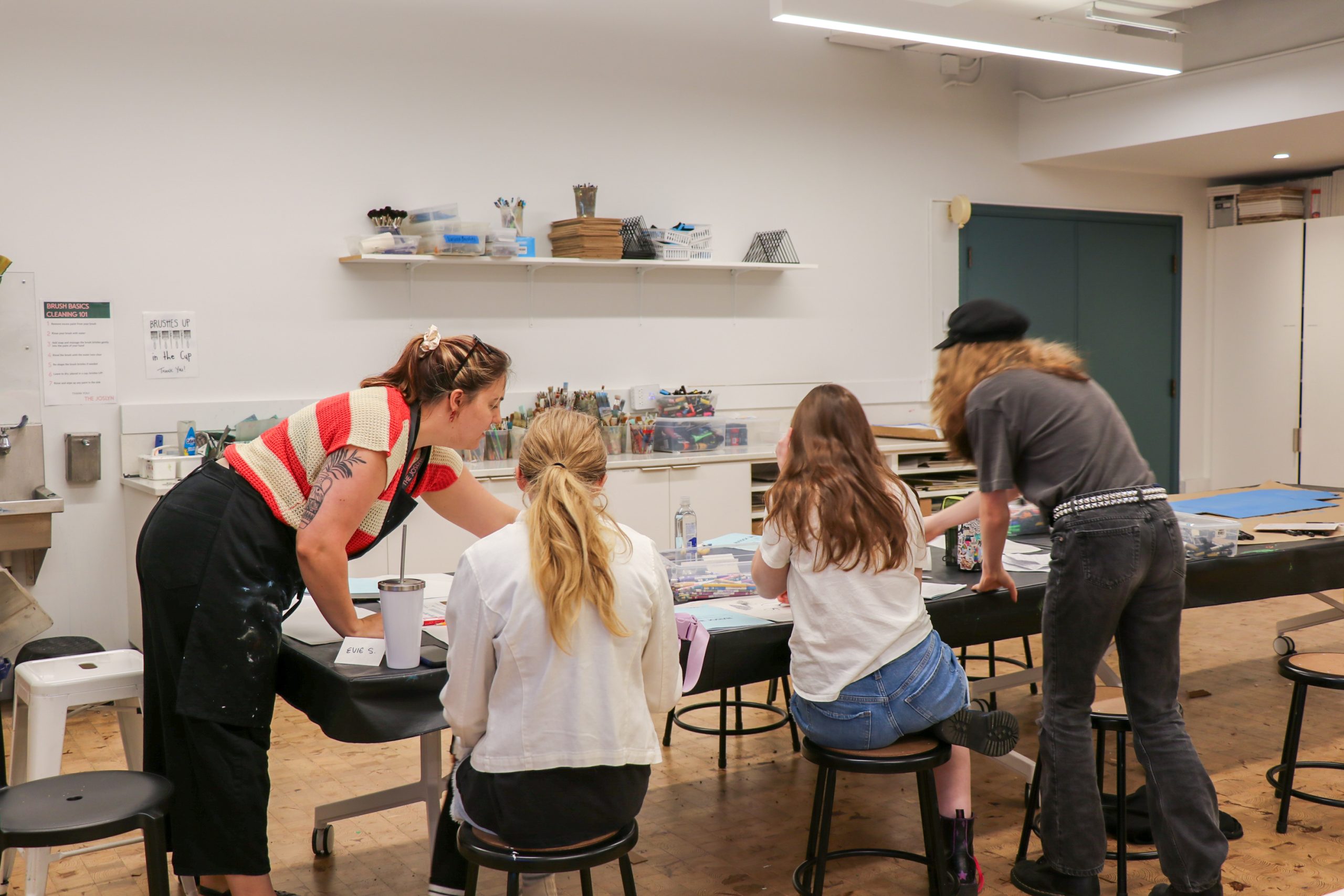 A woman helps three girls with art projects at a table in a classroom, with art supplies and shelves in the background.