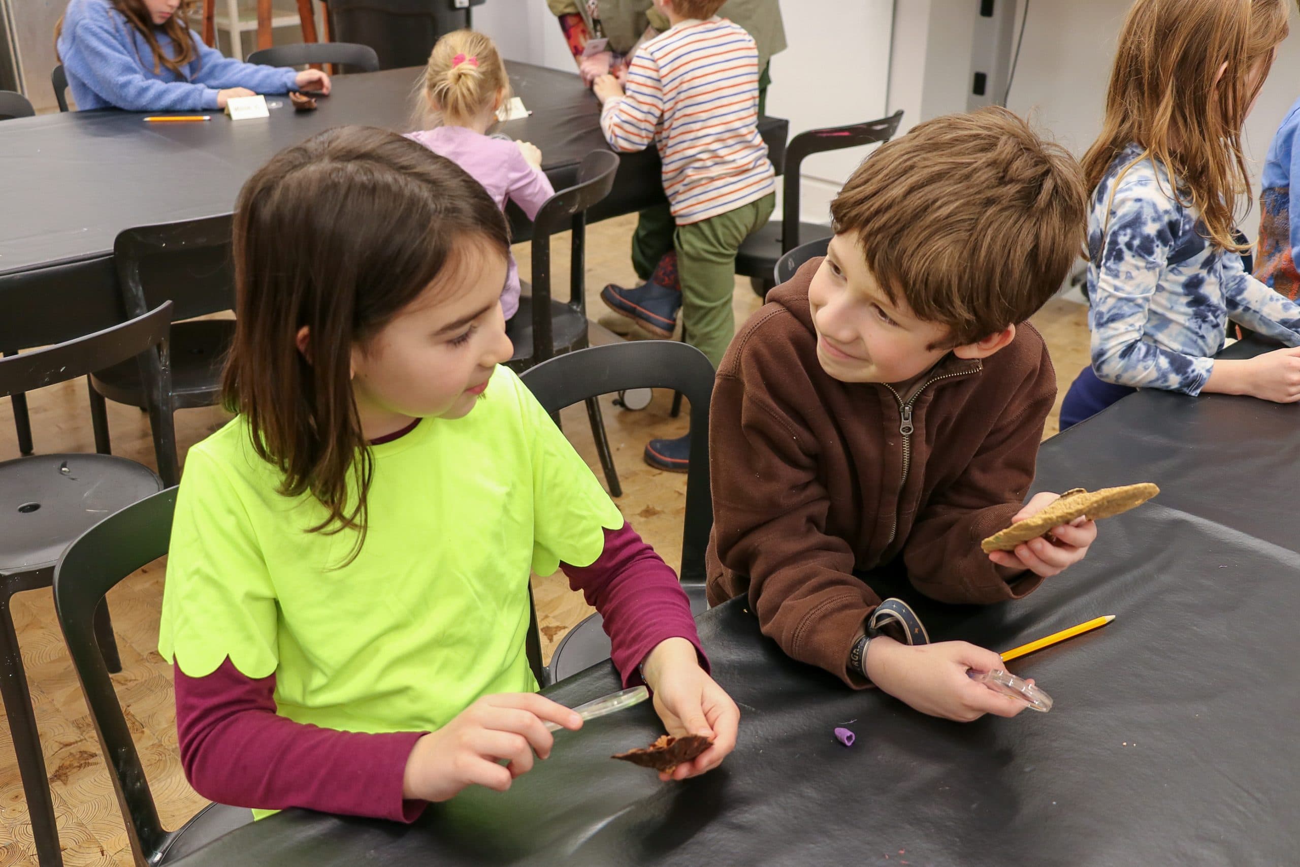 Two children sit at a table, looking at each other while examining objects with magnifying glasses.