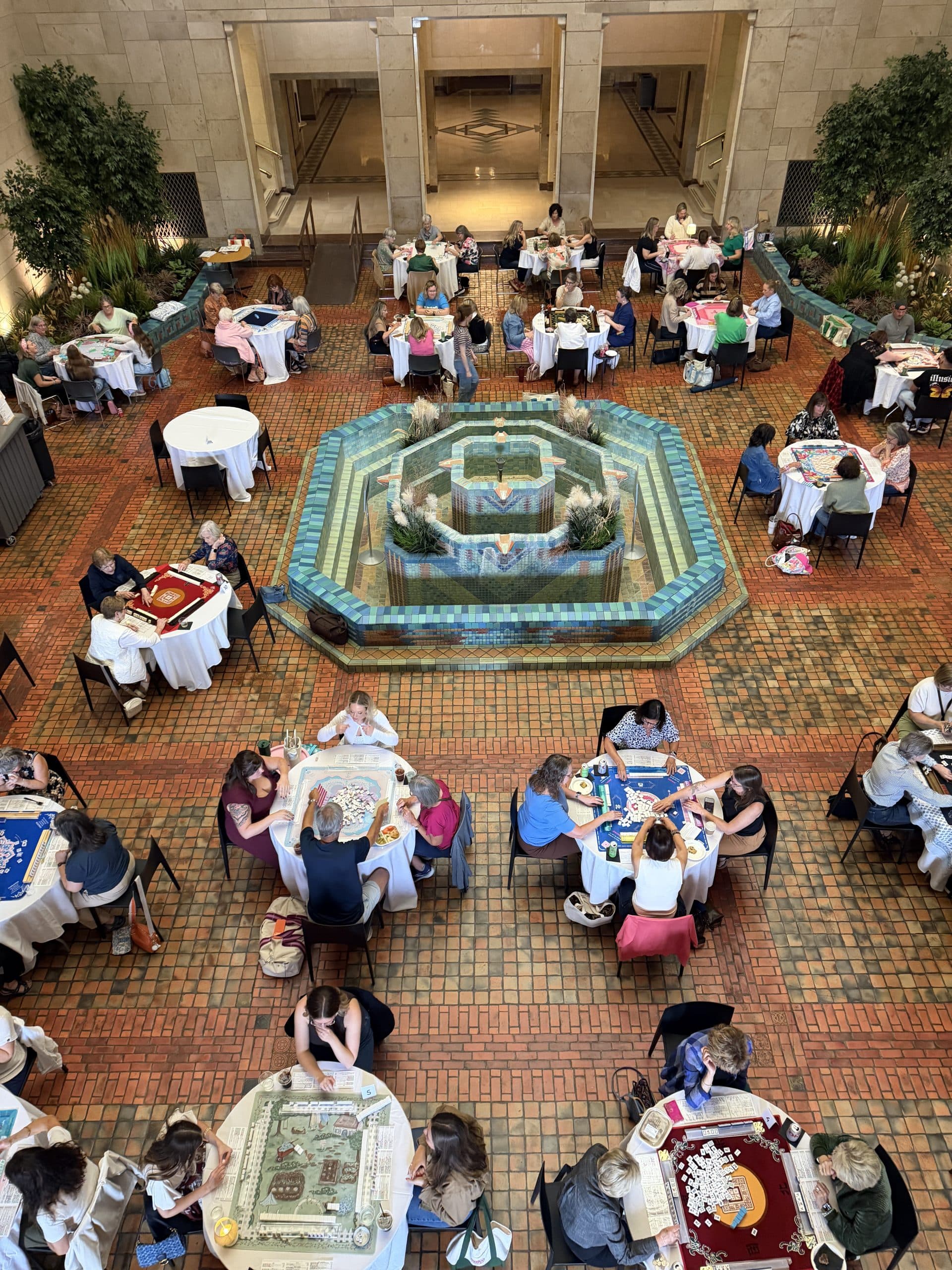 Overhead view of a large room with a tiled fountain in the center, where many people are seated at round tables playing board games.
