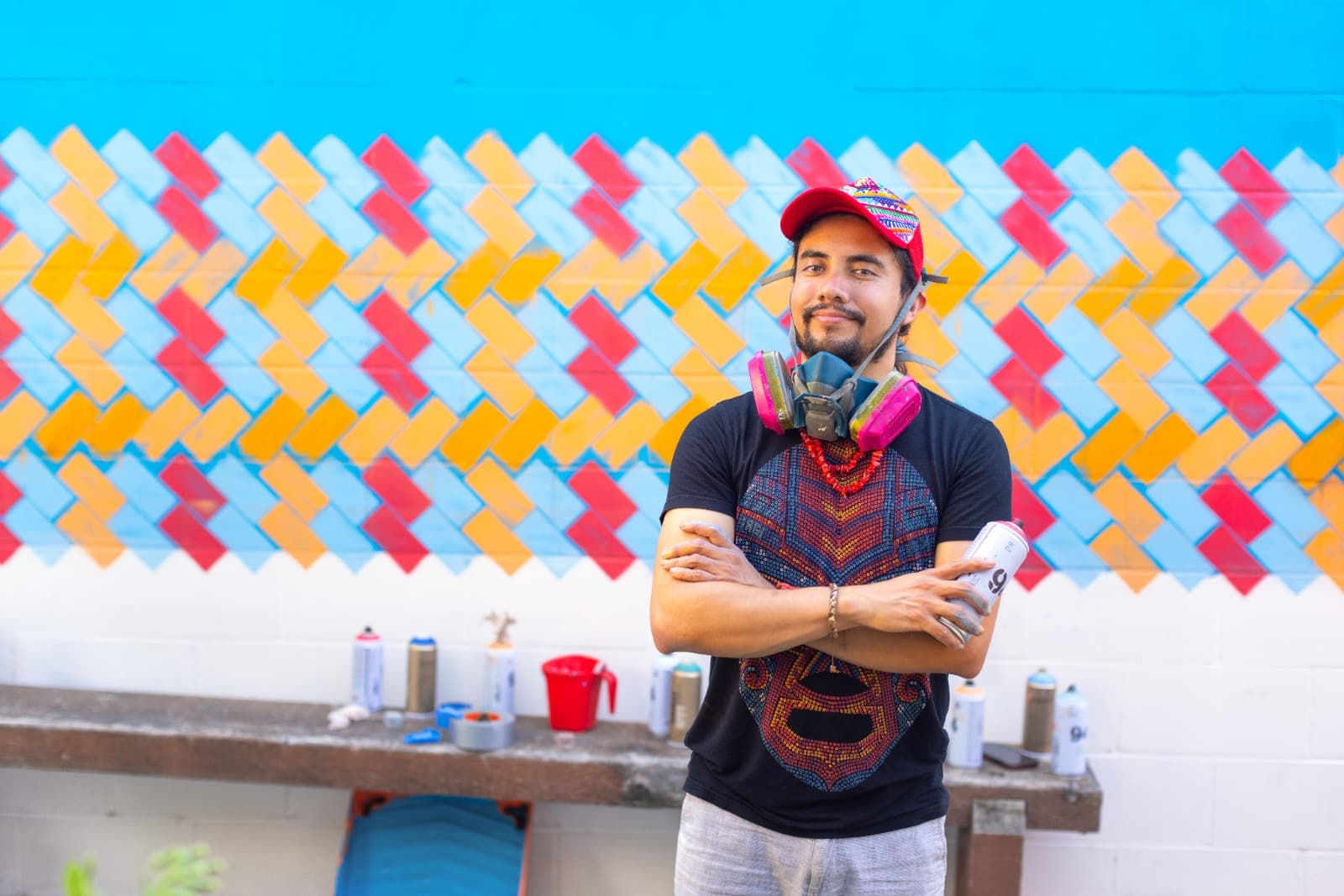 A man in a red cap and black shirt stands in front of a colorful geometric mural, arms crossed, holding a spray paint can with a respirator mask around his neck. Paint cans and supplies are on a bench behind him.