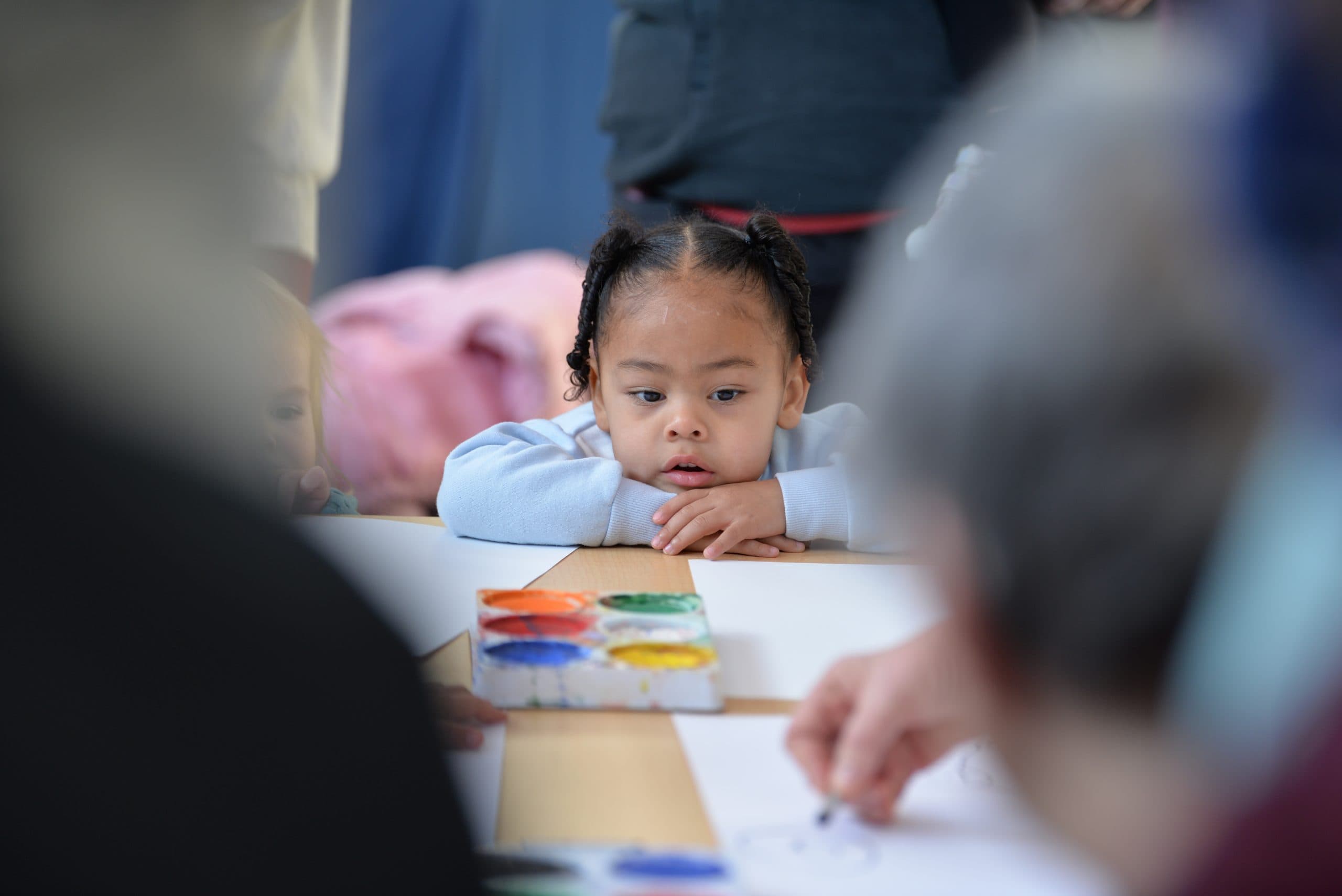 A young child with two braided pigtails sits at a table, leaning forward with their arms resting on the table. They have a focused expression while observing an adult's hands drawing on a piece of paper. Watercolor paints are placed on the table nearby.