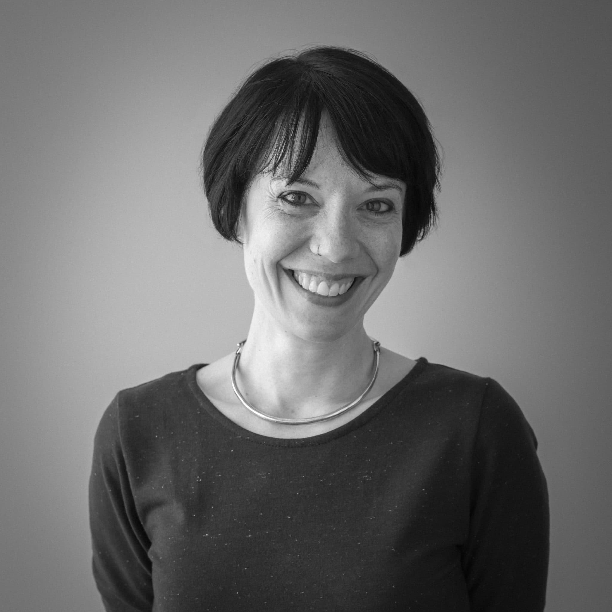 A black and white portrait of a woman with short dark hair, smiling. She is wearing a dark long-sleeved top and a metallic necklace, standing against a plain background.