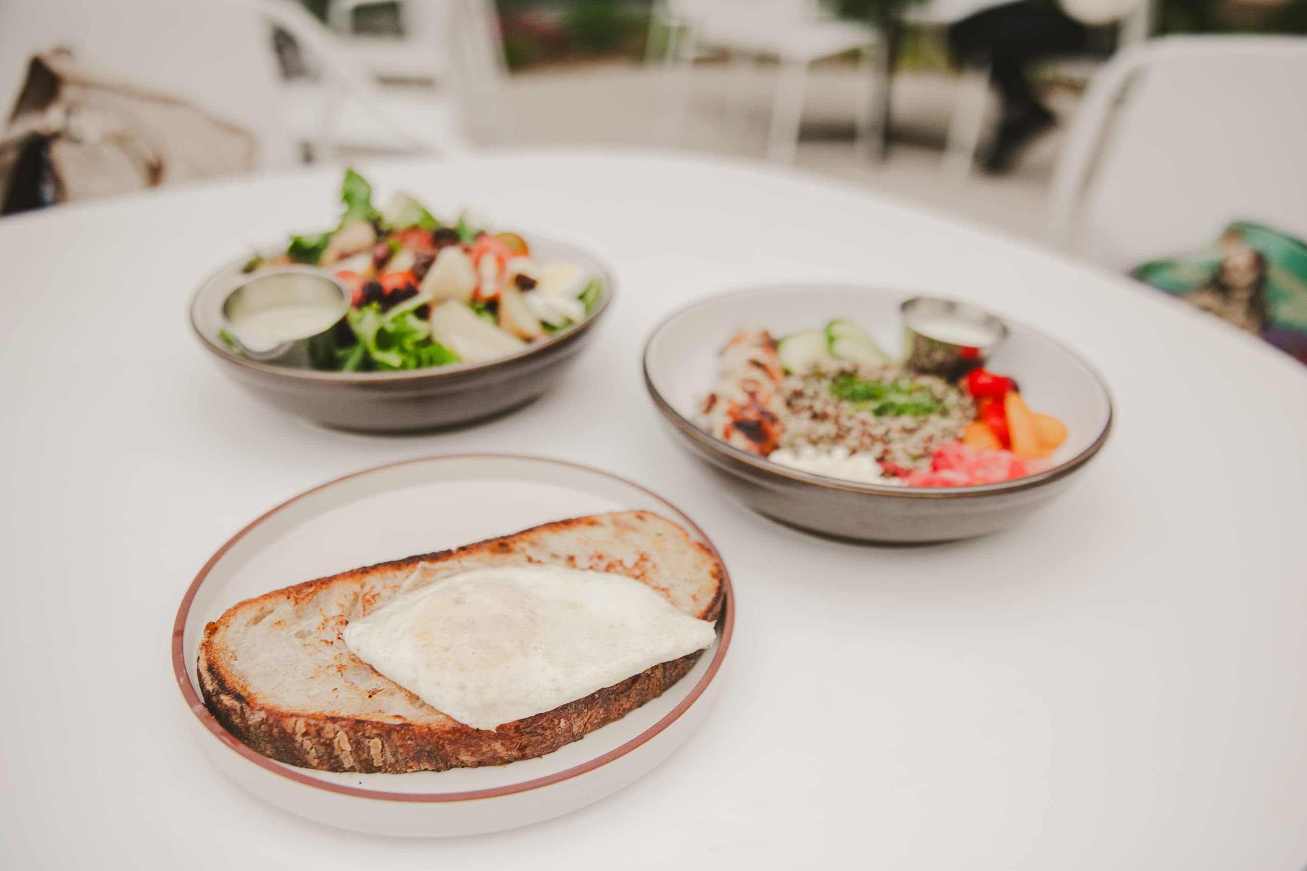 A table setting with a plate of toasted bread topped with a fried egg in the foreground. In the background, there are two bowls, one containing a colorful salad and the other a mix of grains, vegetables, and a sauce.