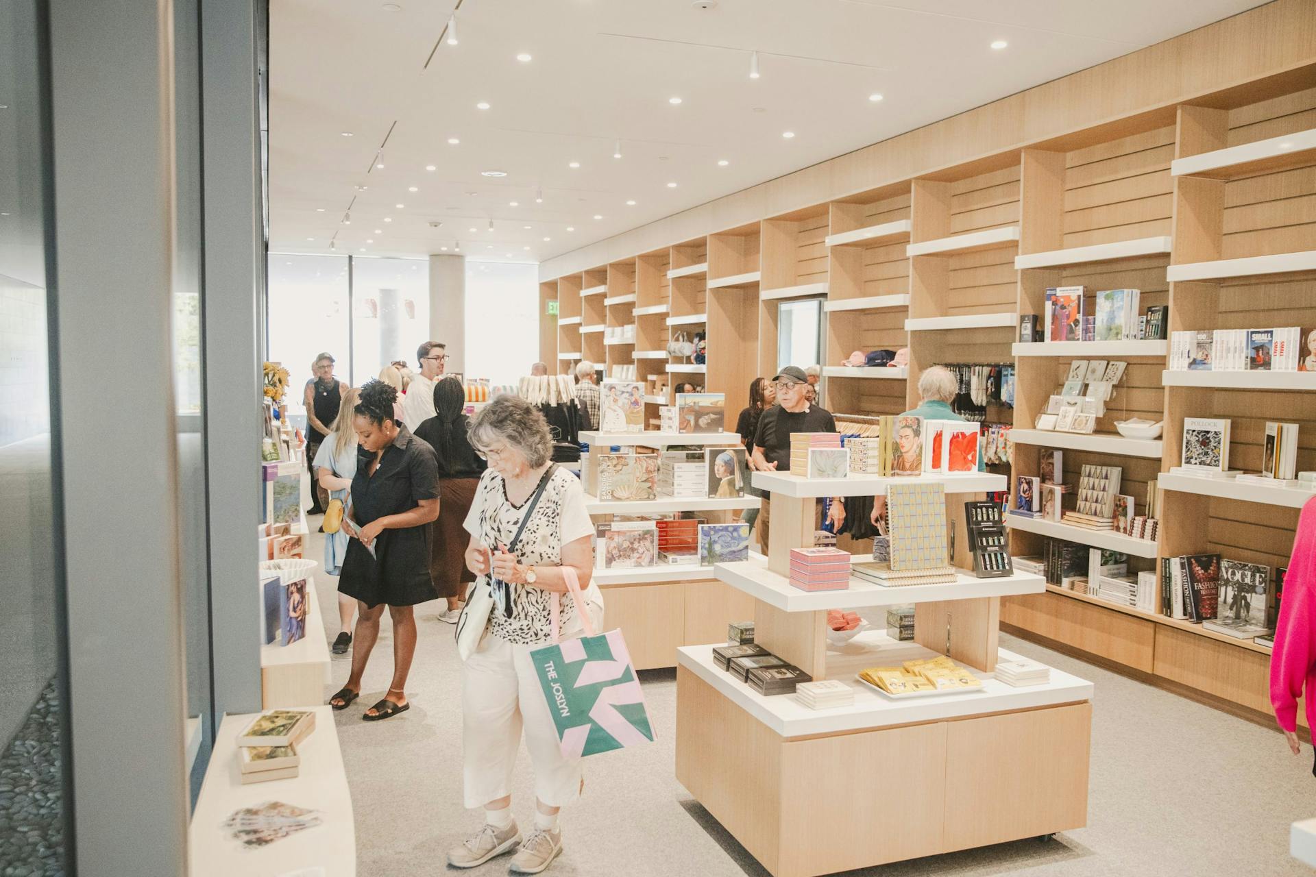 A brightly lit bookstore with wooden shelves filled with books and merchandise. Several people browse the aisles and examine items. In the foreground, a woman wearing white holds a shopping bag, and other customers are visible in the background.