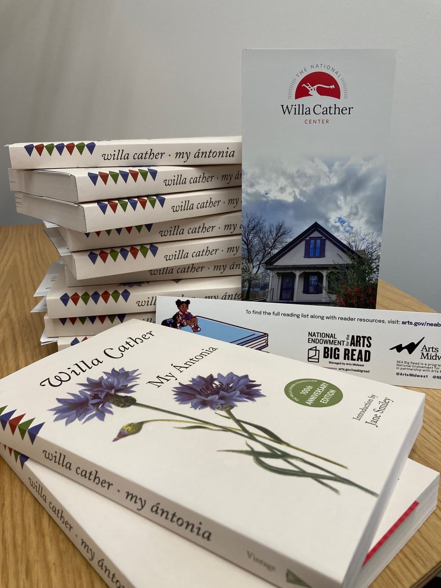 A stack of "My Ántonia" books by Willa Cather sits on a wooden table. A brochure for the Willa Cather Center is standing beside them. The book cover features flowers, and the brochure displays a house and trees.