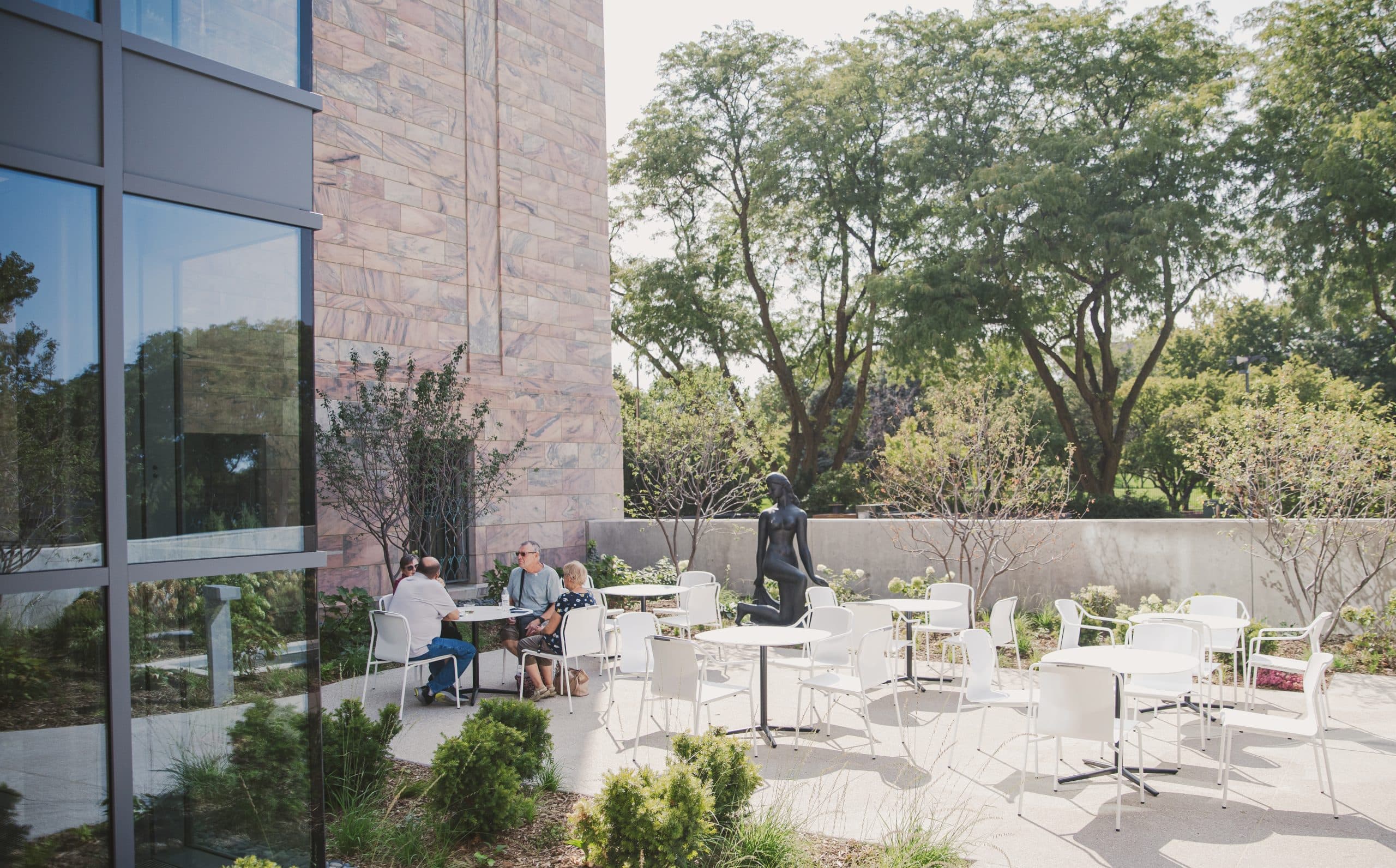 A group of people sitting at a table on an outdoor patio, surrounded by empty white tables and chairs. The patio is adjacent to a building with large glass windows. In the background, there are leafy green trees and a stone wall. The atmosphere is sunny and serene.