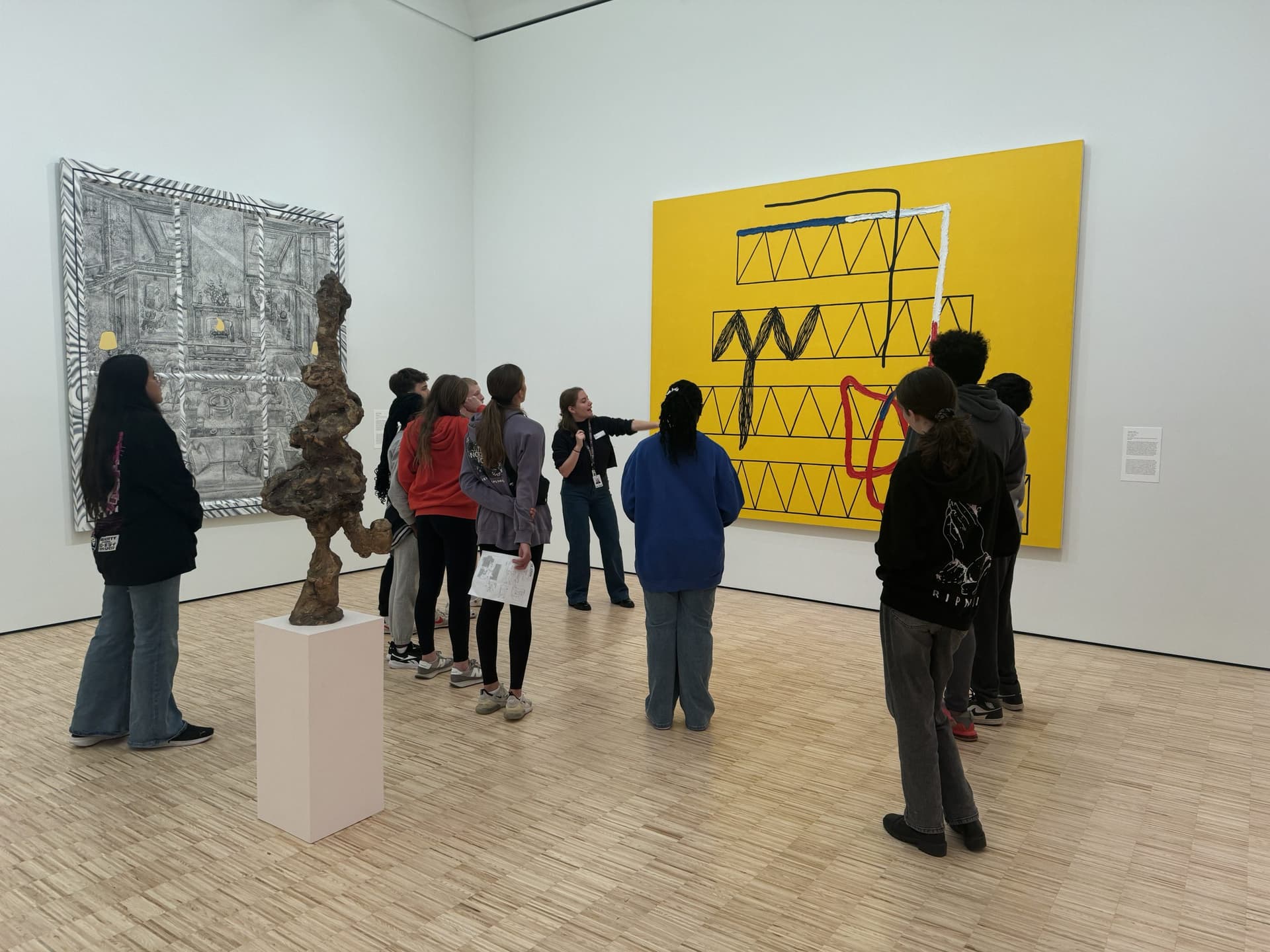 A group of people in a gallery observes two large abstract paintings on the walls and a unique sculpture on a pedestal. The room has light wooden floors and white walls, creating a modern, spacious atmosphere.