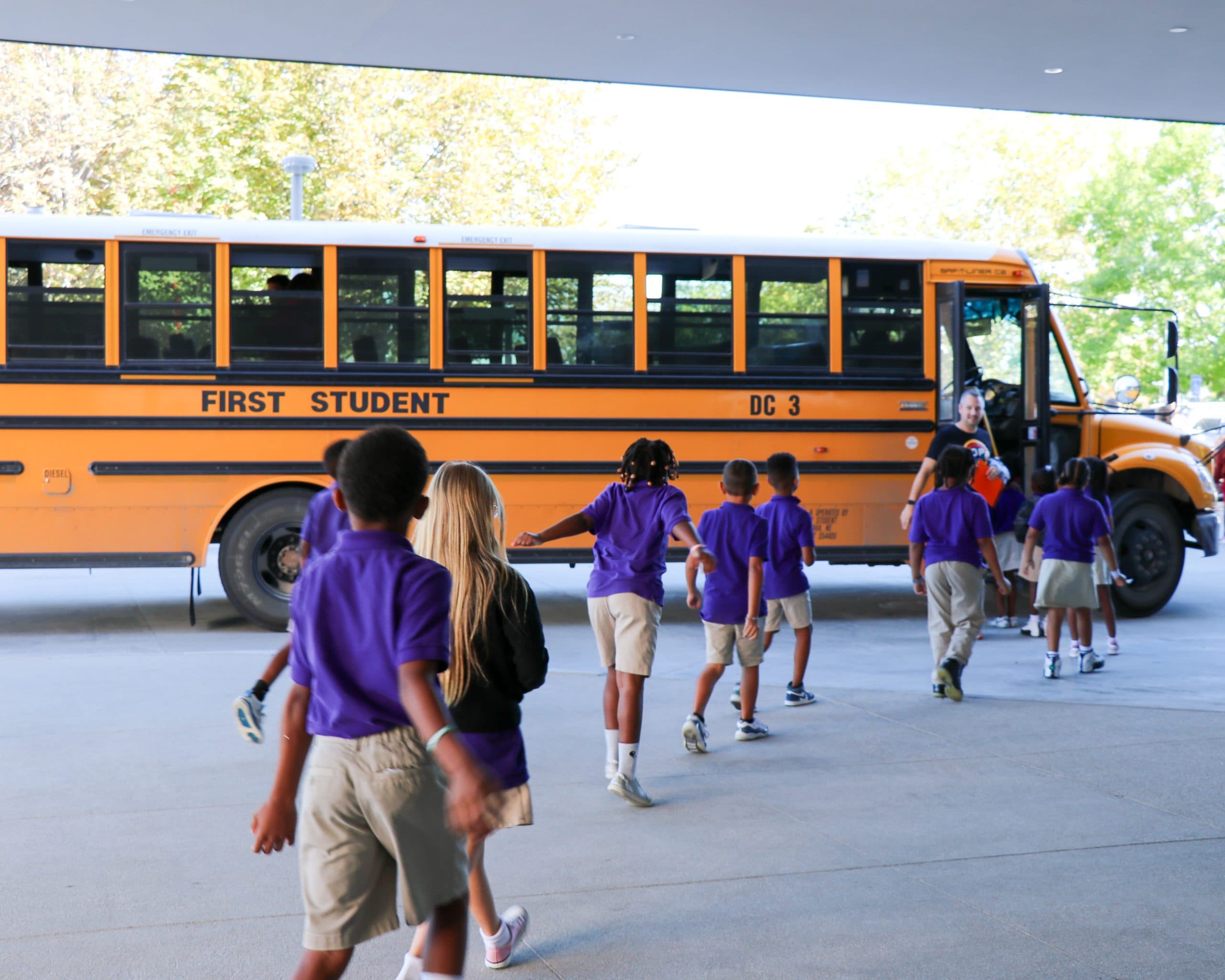 Children in school uniforms are walking in a line towards a yellow school bus marked "First Student." The background shows trees and a bright sky. The scene depicts a school environment with children boarding the bus.