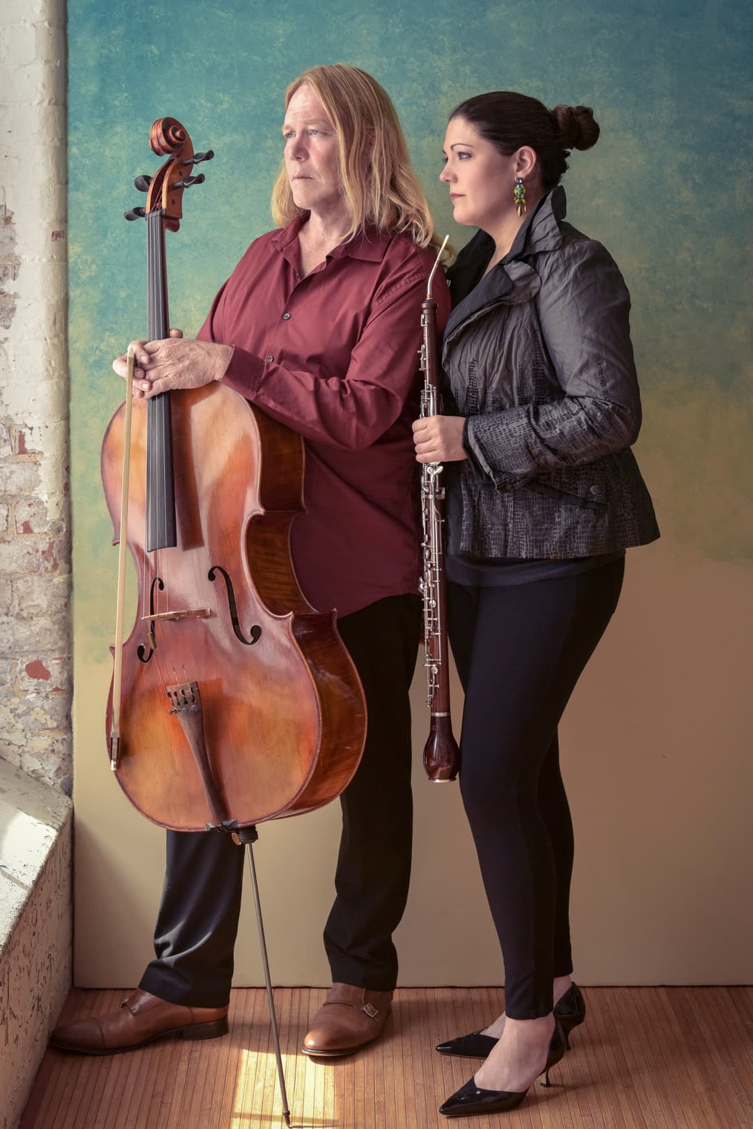 A man holding a cello stands next to a woman holding an oboe. They are posing in front of a textured, blue-green wall. The man has long blond hair and wears a red shirt. The woman has dark hair tied back and wears a black jacket and pants.